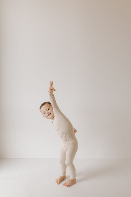 A child in cozy Bamboo Two Piece Pajamas by forever french baby poses joyfully with one arm raised in a minimalist, light-toned room. The hypo-allergenic comfort of light-colored bamboo enhances the playfulness of their smile and pose.