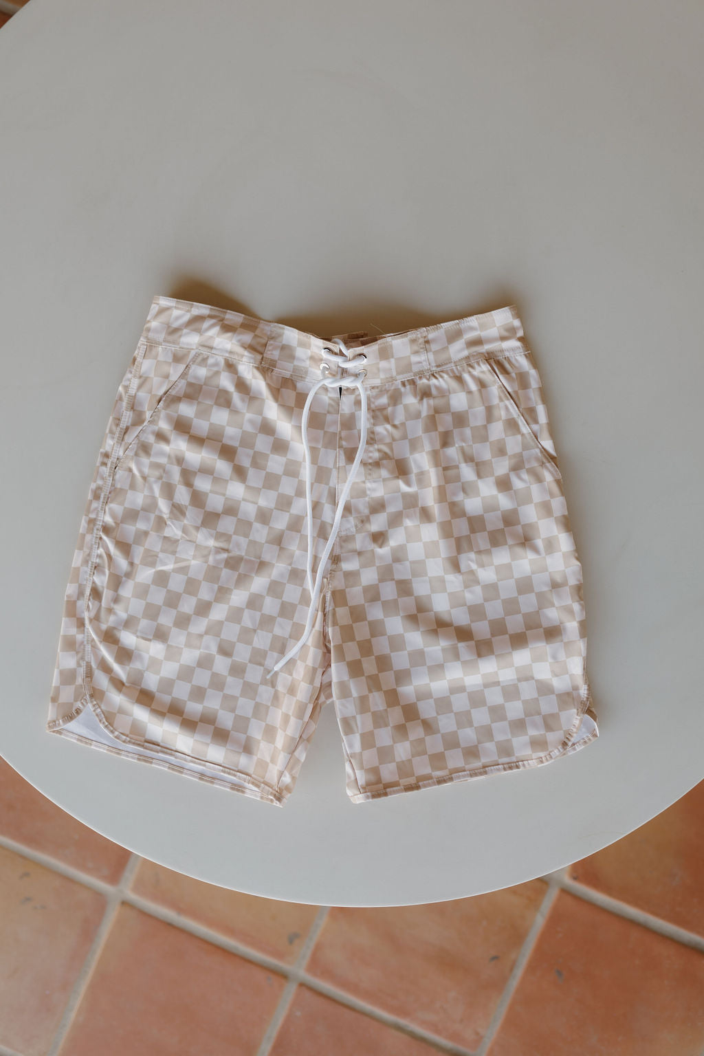 Men's Boardshort | Coastline from forever french baby in beige and white checkered pattern with a drawstring waistband displayed on a round white table atop terracotta tiles, embodying the essence of Forever French Baby style.