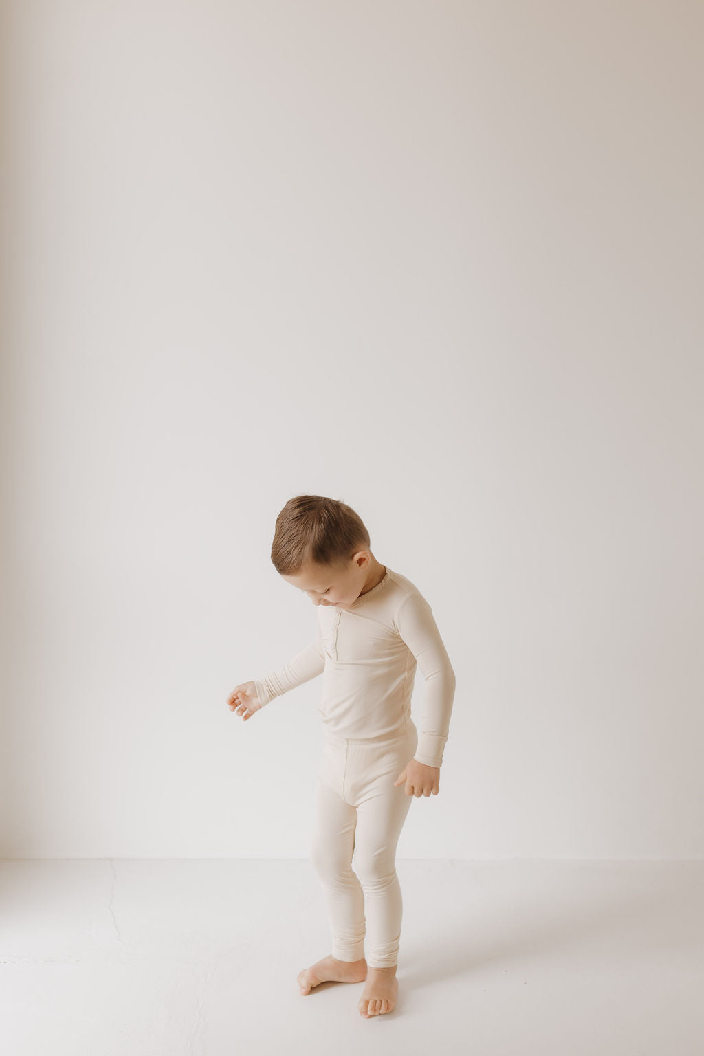 A child stands barefoot on a white surface in "Bone" Bamboo Two Piece Pajamas by forever french baby, gazing slightly downward. The off-white background enhances the minimalist and serene setting. These hypo-allergenic pajamas are ideal for delicate skin.