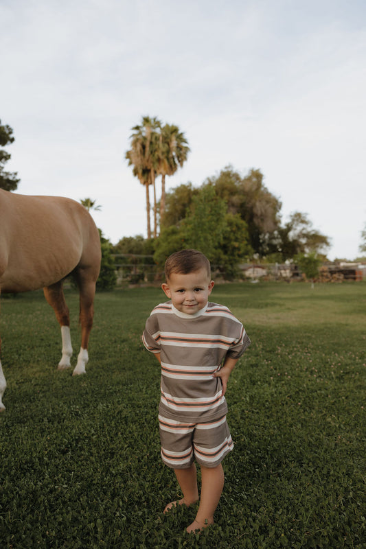 A young child wears the forever french baby Child Short Set | Sedona Stripe, standing barefoot on grass with hands on hips and smiling. A horse appears to the left, with palm trees and green foliage beneath a partly cloudy sky.