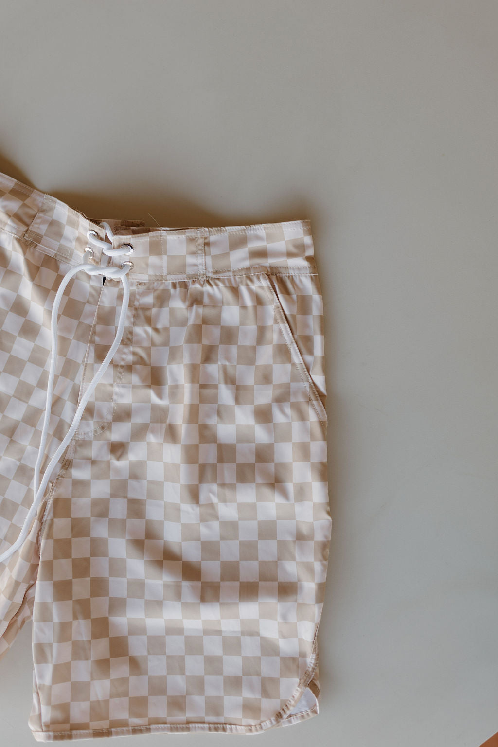 The men's boardshort from forever french baby's Coastline Collection comes in beige and white checks with a white drawstring waistband. Displayed on a neutral background, these shorts have side pockets and the fabric is presented flat.