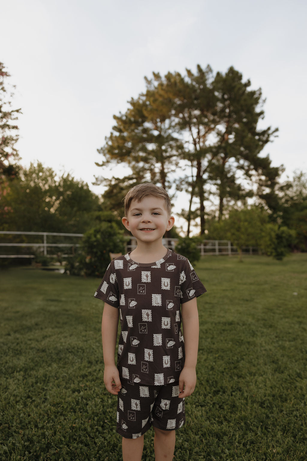 A young boy with short brown hair smiles on a grassy lawn, wearing the Bamboo Short Set by forever french baby, featuring white geometric patterns. Tall green trees and a fence are in the background under a partly cloudy sky.