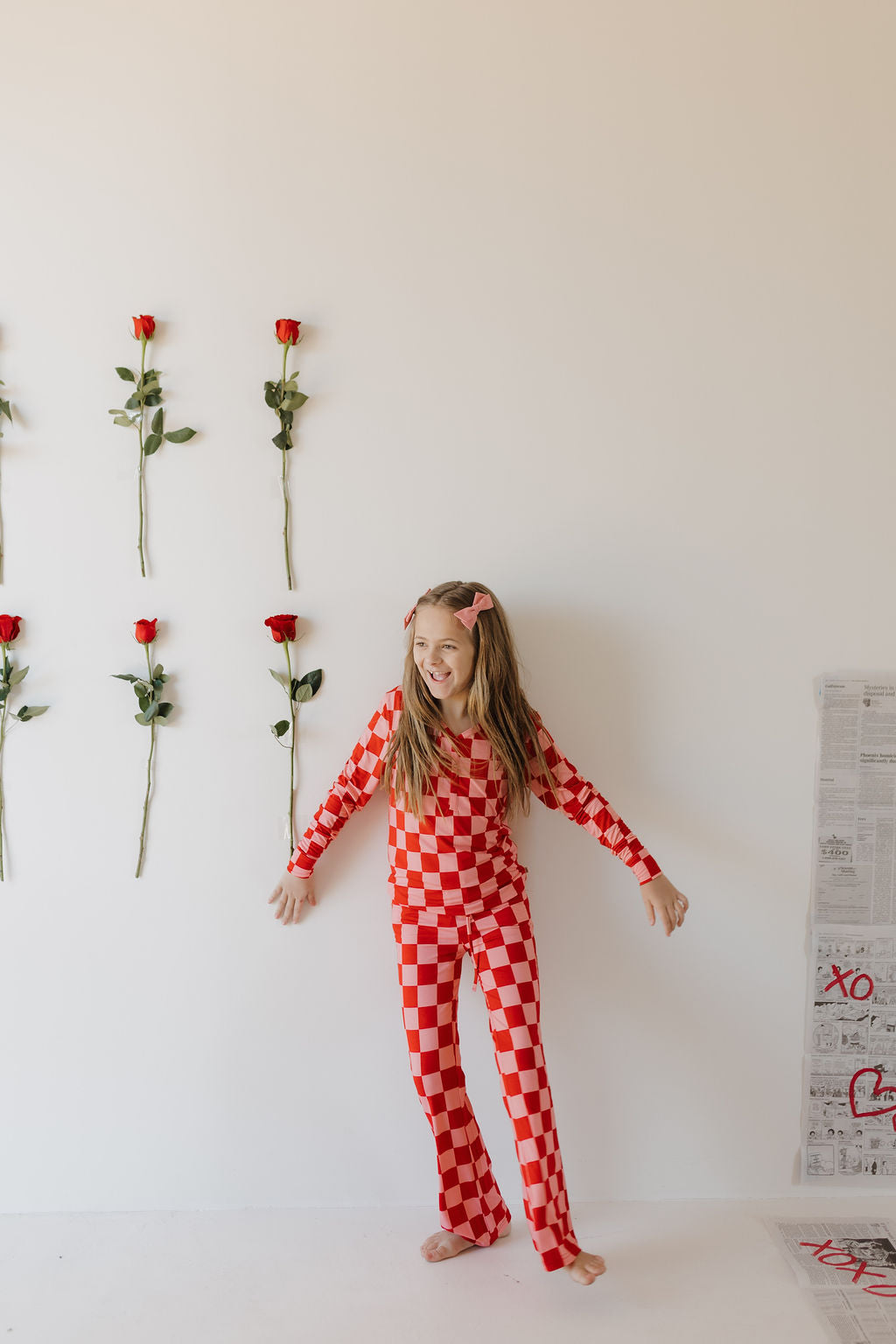 Against a white wall adorned with roses, a young girl with long hair smiles in her red and white Youth Flare Bamboo Pajamas by forever french baby. Newspapers are neatly stacked on the floor beside her.