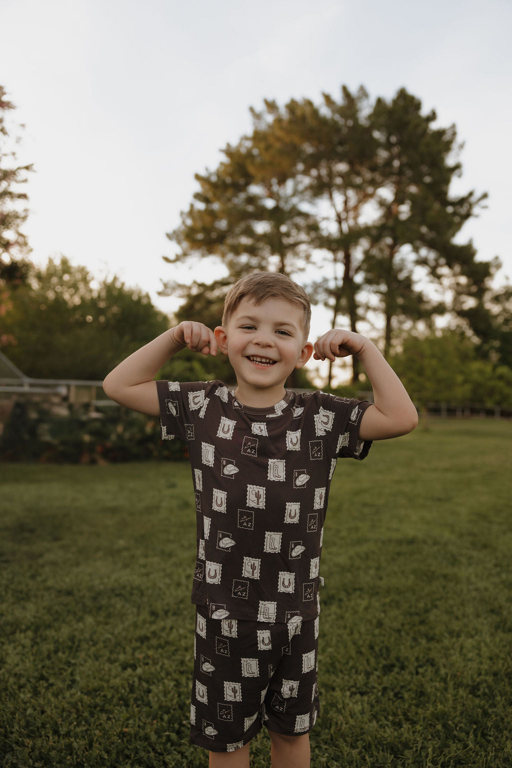 A young boy stands on grass, smiling and flexing in the comfy hypoallergenic Bamboo Short Set by forever french baby. The matching t-shirt and shorts feature a fun pattern. Trees and a bright sky complete the scene.