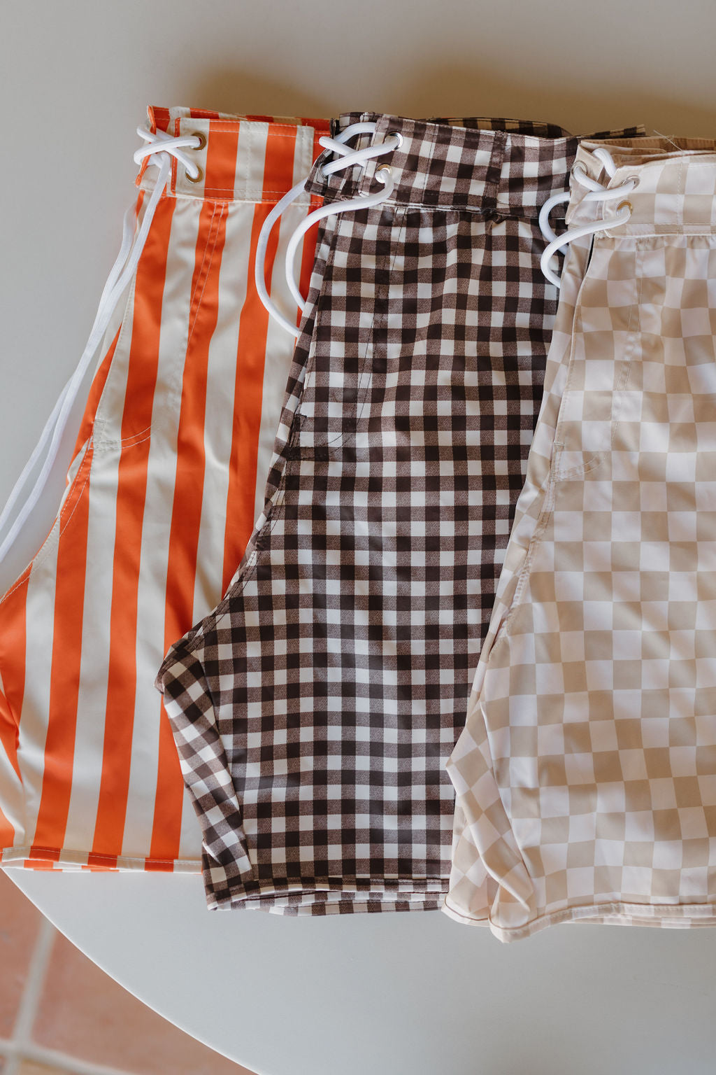 Three pairs of high-waisted Men's Boardshorts from forever french baby's Poolside collection laid out: one with red and white vertical stripes, one with a black and white checkered pattern, and one featuring beige and white checks.