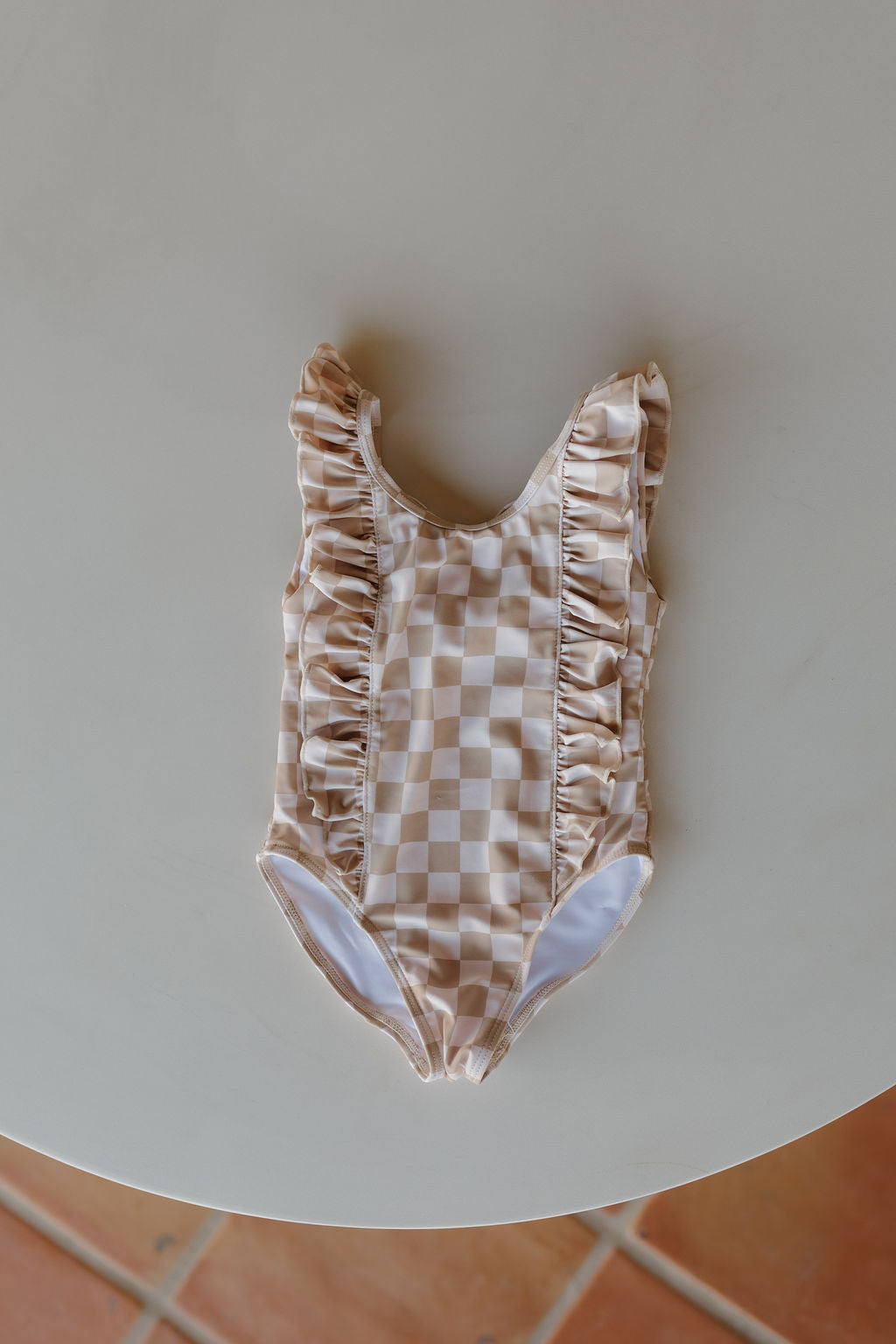 From the Coastline Collection by forever French baby, this timeless Girls Sleeveless Swimsuit for toddlers showcases a charming beige and white checkered pattern with ruffle details, beautifully displayed on a light surface.