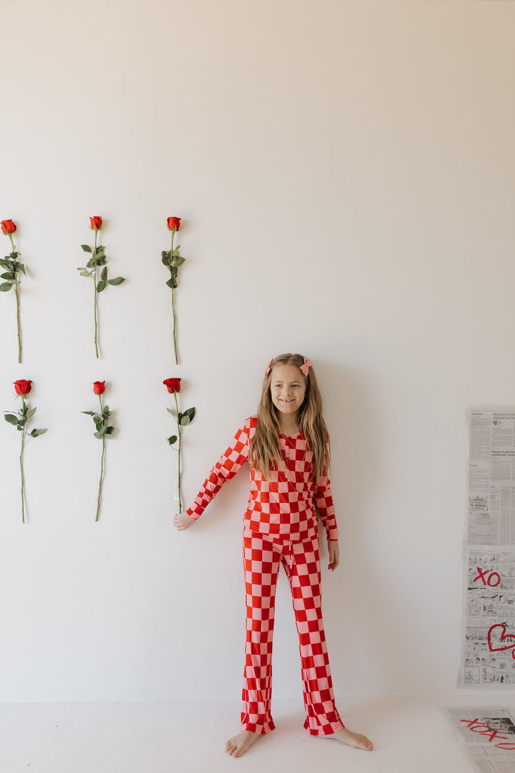 A young girl in "Youth Flare Bamboo Pajamas | XOXO" by forever french baby stands barefoot against a white wall, holding a red rose. Five vertical roses adorn the wall beside her, while newspapers are stacked on the floor to her right.