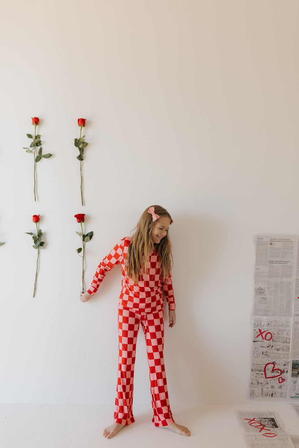 A girl in forever french baby Youth Flare Bamboo Pajamas | XOXO, with red and white checks, stands by a white wall holding a red rose. More roses are attached beside her. Her long hair is adorned with a pink bow, while newspapers with doodles scatter the floor. The breathable fabric enhances her charm.