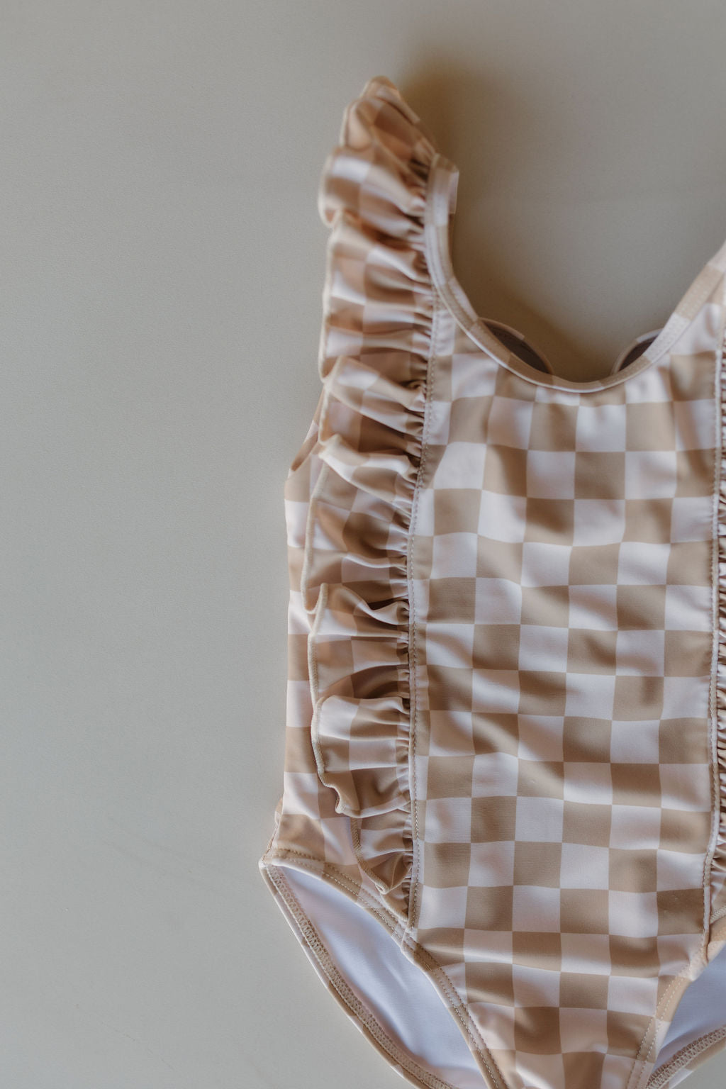 The Girls Sleeveless Swimsuit from the Coastline Collection by Forever French Baby features a beige and white checkerboard pattern with elegant ruffles along the neckline and shoulders, capturing timeless charm as it lays flat on display.