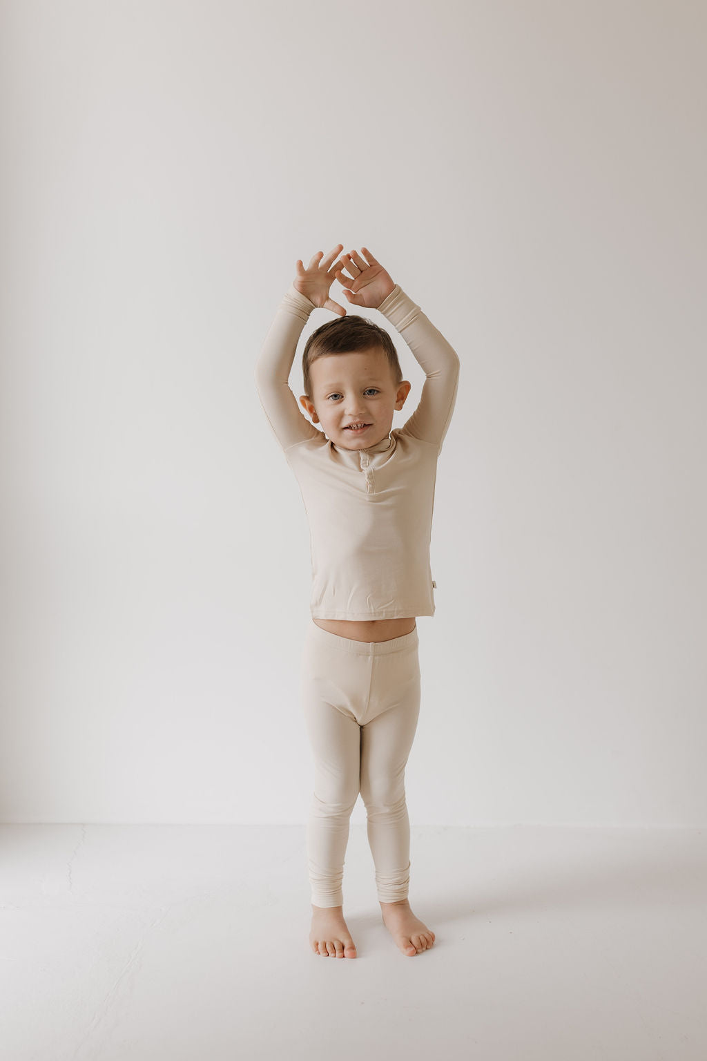 A small child stands barefoot on a white floor, wearing forever french baby's Bamboo Two Piece Pajamas in Bone with arms raised above their head. The breathable sleepwear complements the plain light gray wall behind.