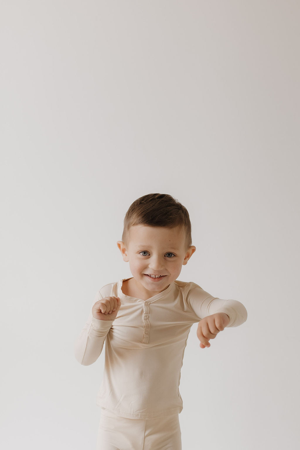 A young child with short brown hair poses playfully against a light gray background, wearing cozy Bamboo Two Piece Pajamas in Bone by forever french baby, promising comfort and a smile.