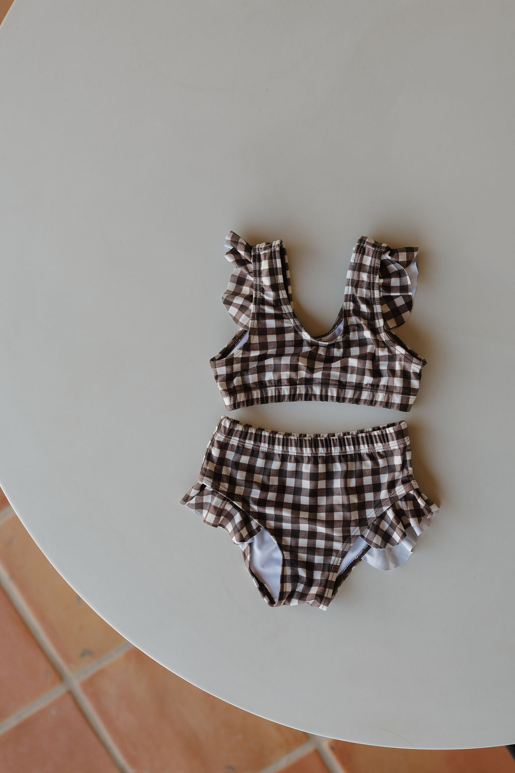 From forever french baby's Chocolate Gingham Collection, the Girls Two Piece Swimsuit in brown and white checkered pattern, featuring ruffled straps and waistband, is showcased on a round white surface.