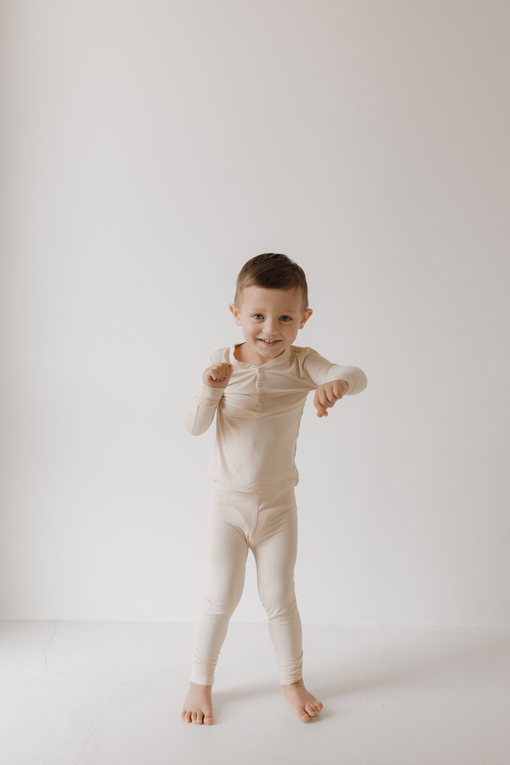 A young child with short brown hair stands barefoot against a plain white background, wearing forever french baby's cozy and hypo-allergenic Bamboo Two Piece Pajamas in Bone. The child smiles while striking a playful pose.