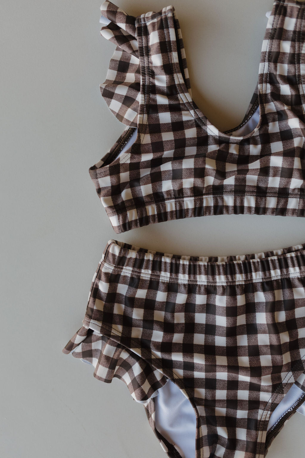 Part of the forever french baby collection, the Girls Two Piece Swimsuit in Chocolate Gingham showcases a brown and white pattern. It includes a top with ruffled shoulder straps and high-waisted bottoms adorned with side ruffles, displayed elegantly on a light surface.