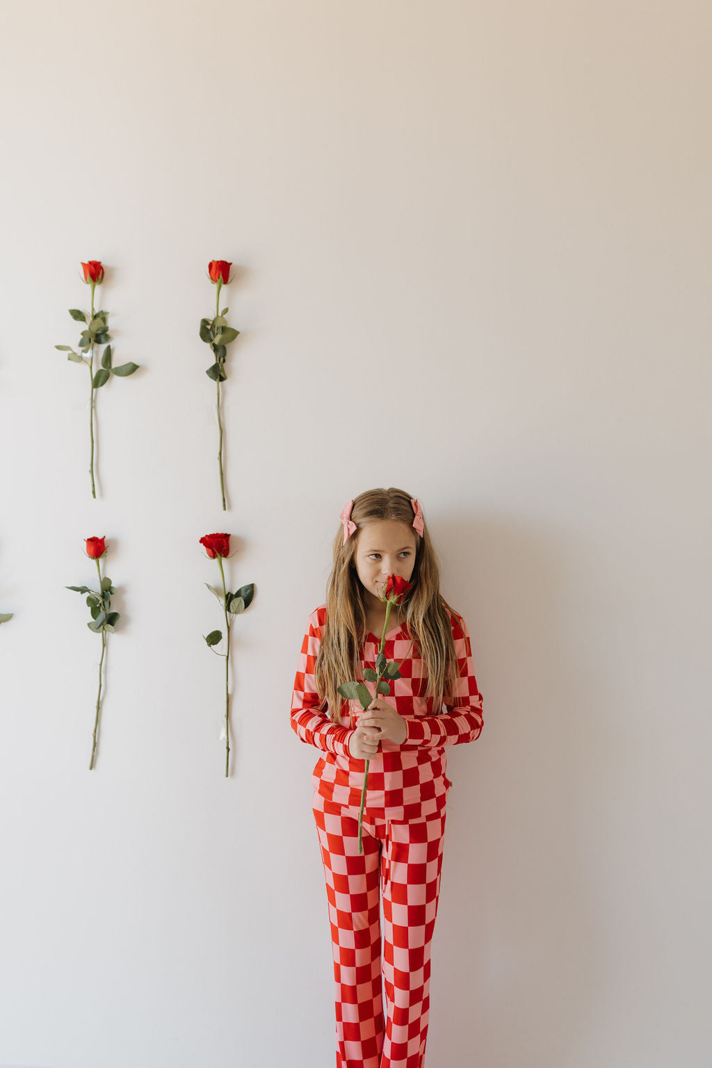 A child with long hair, adorned with pink clips, stands against a white wall in forever french baby Youth Flare Bamboo Pajamas | XOXO. They hold a red rose and six roses are taped vertically to the wall in two symmetrical rows.