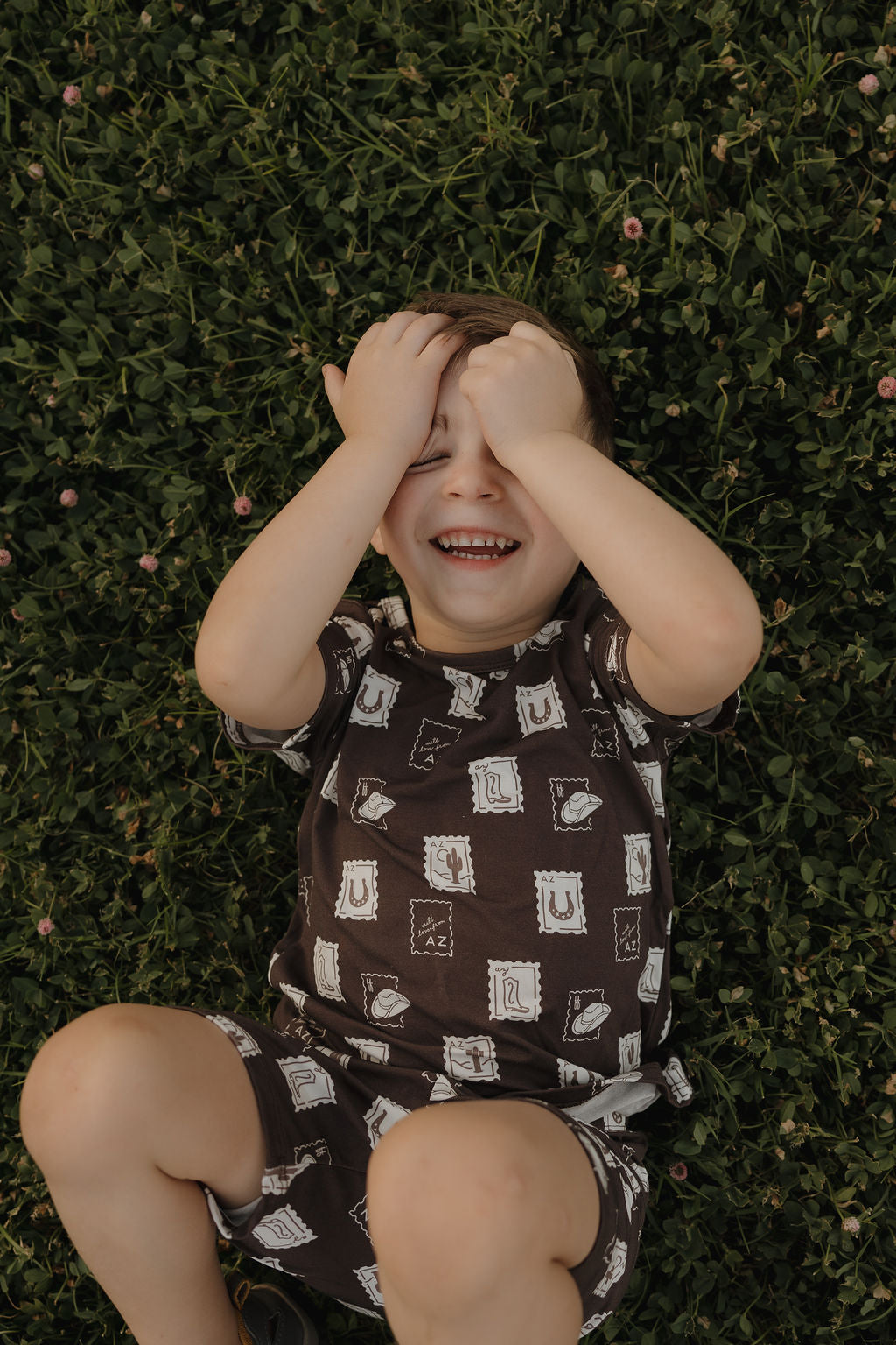 A young child wearing the forever french baby Bamboo Short Set | With Love from AZ lies on green grass, smiling and covering their eyes, surrounded by clover flowers.
