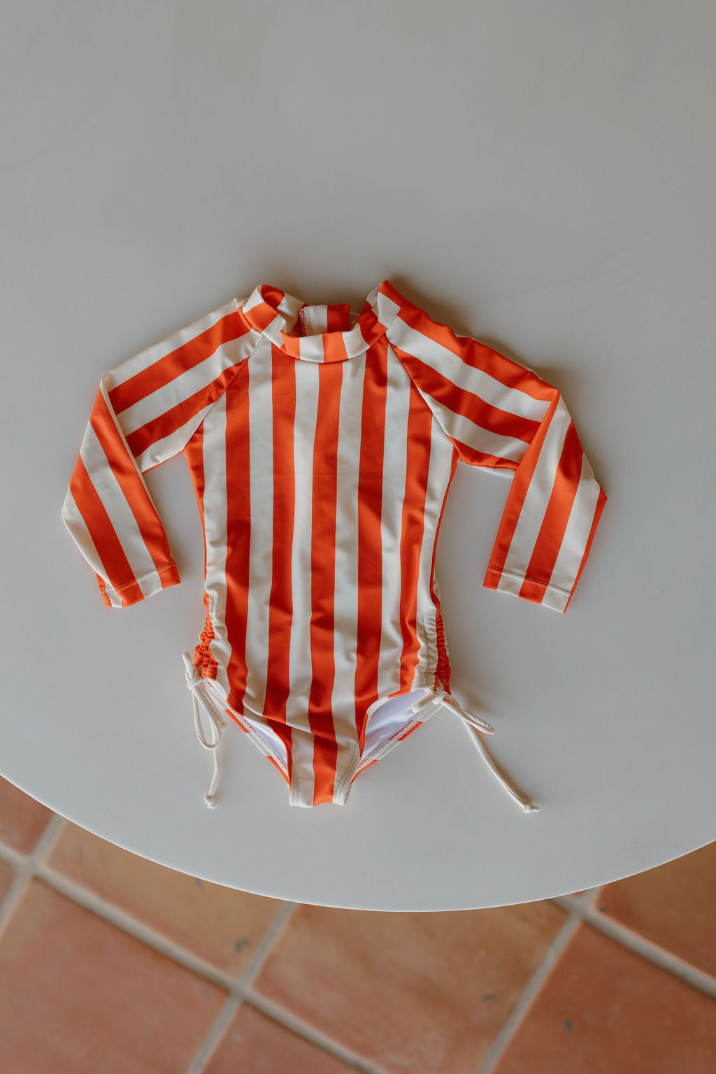 The forever french baby Girls Long Sleeve Swimsuit from the Poolside Collection features vertical orange and white stripes with side drawstrings. It is stylishly positioned on a circular white tabletop, complemented by glimpses of a tiled floor.