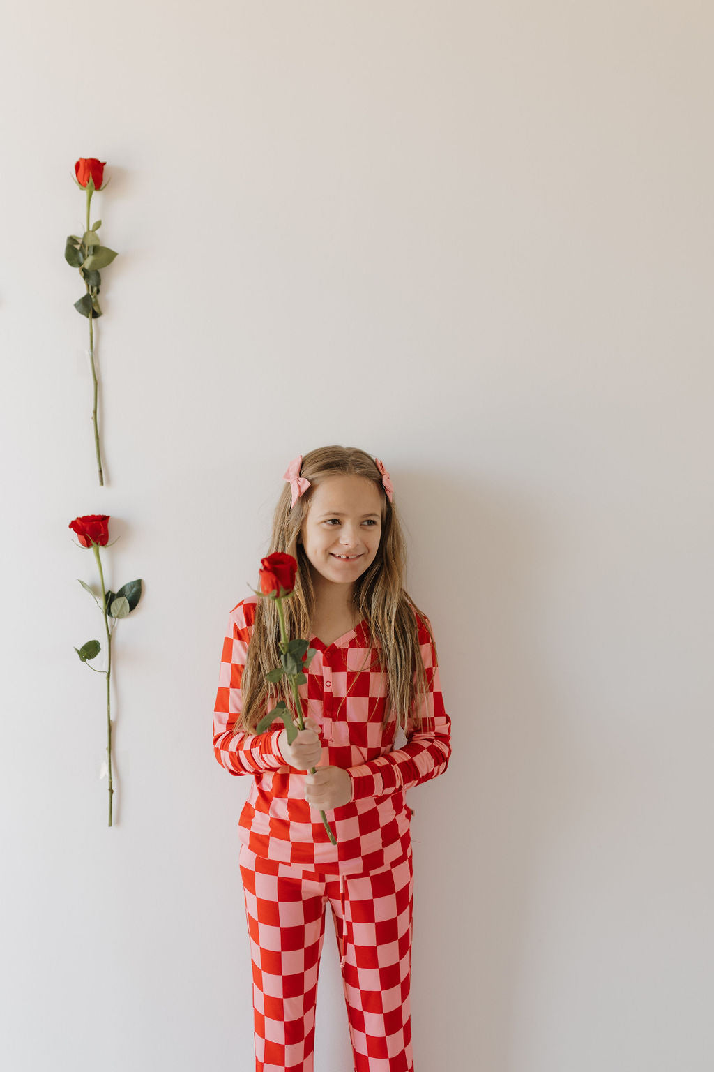 A young girl in red and white Youth Flare Bamboo Pajamas from forever french baby smiles while holding a red rose. She stands against a white wall with three vertical roses taped to it, her long hair with pink clips enhancing her playful charm.