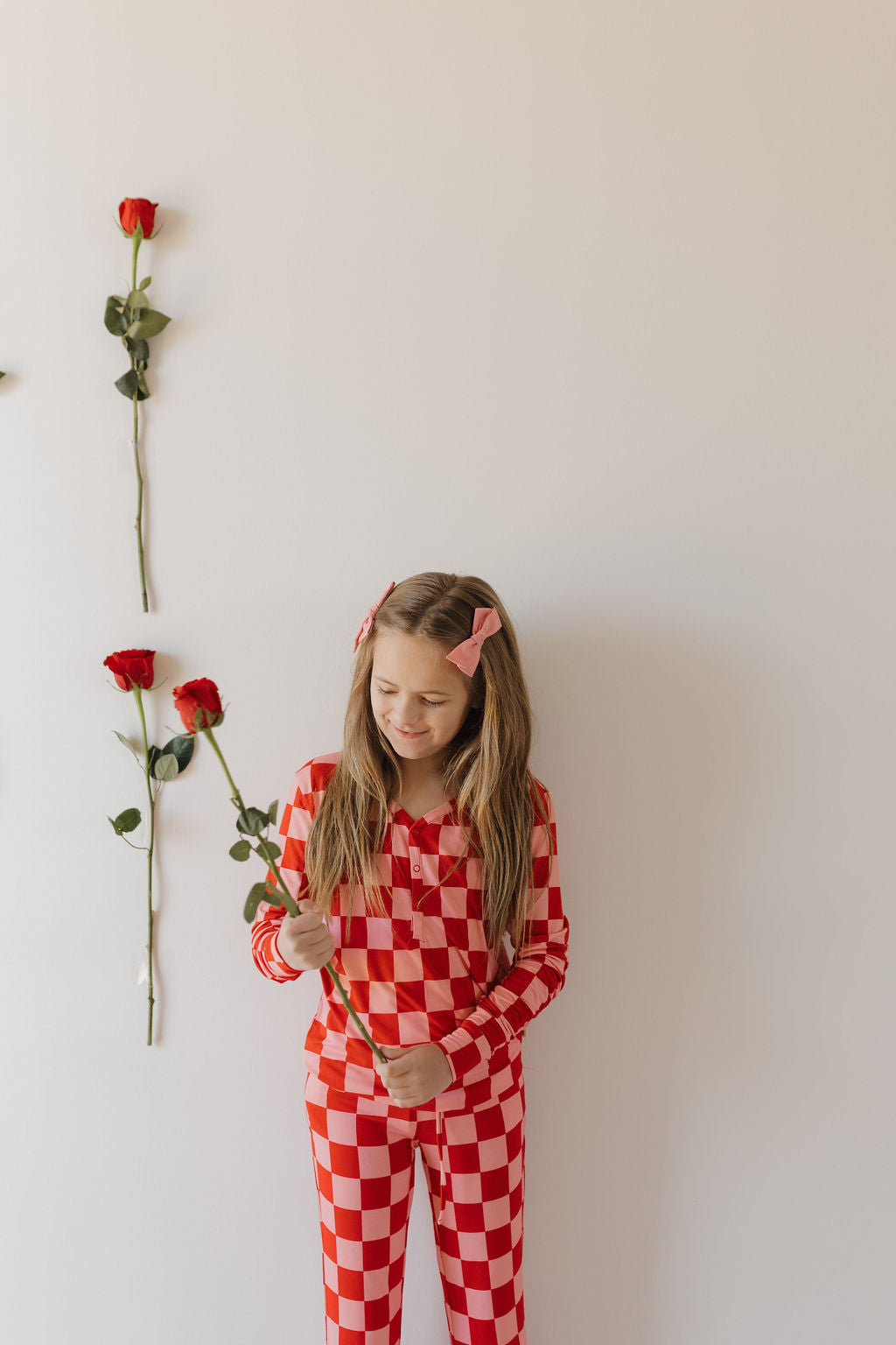 A girl wearing forever french baby's Youth Flare Bamboo Pajamas | XOXO, adorned with red and white checkered fabric, holds a red rose. She stands against a white wall with three roses and her hair is styled with pink bows.