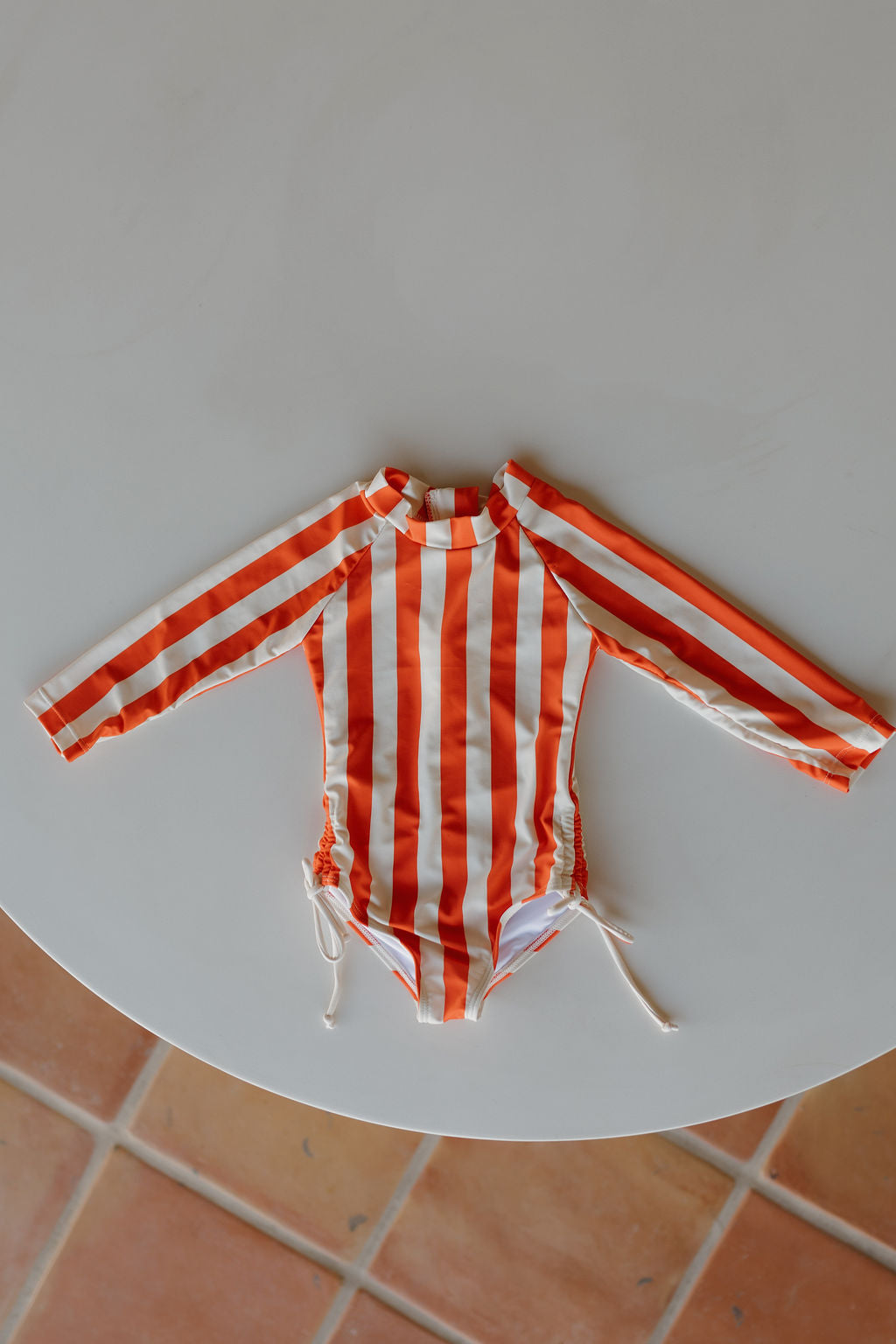 The Poolside Collection by forever french baby features a chic Girls Long Sleeve Swimsuit with orange and white stripes, laid flat on a round white surface. Highlighted by drawstring sides, it is showcased beautifully against a tiled floor backdrop.