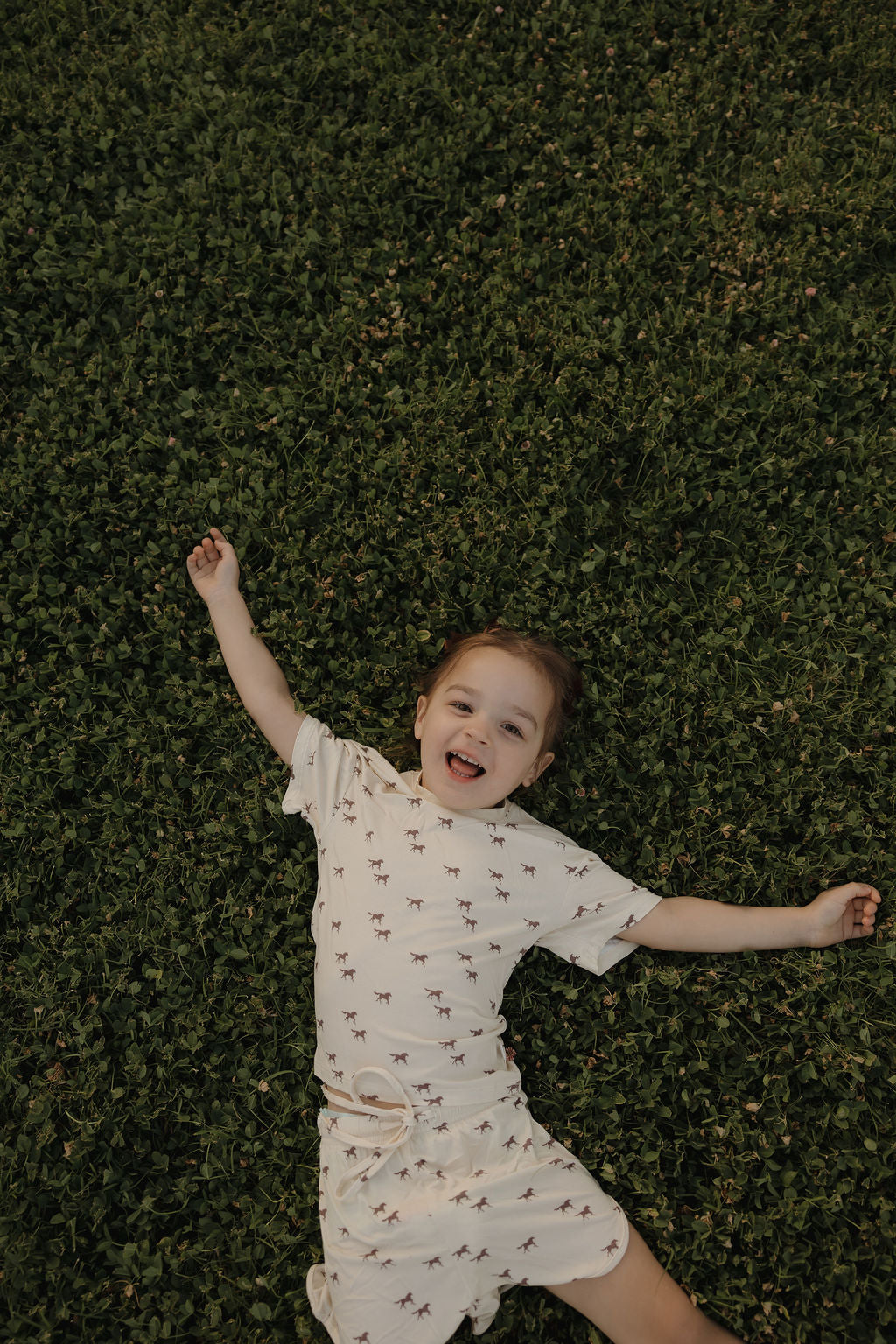 A smiling young child lies back in the grass wearing the forever french baby Girl's Bamboo Short Set | Wild Horses, a soft, hypo-allergenic outfit featuring a playful horse print.