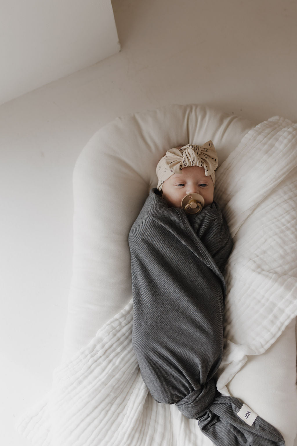 A baby wrapped in the forever french baby Bamboo Swaddle | Shadow rests on a white cushion, with a pacifier and beige headband featuring a bow. The soft lighting enhances this peaceful and cozy scene.