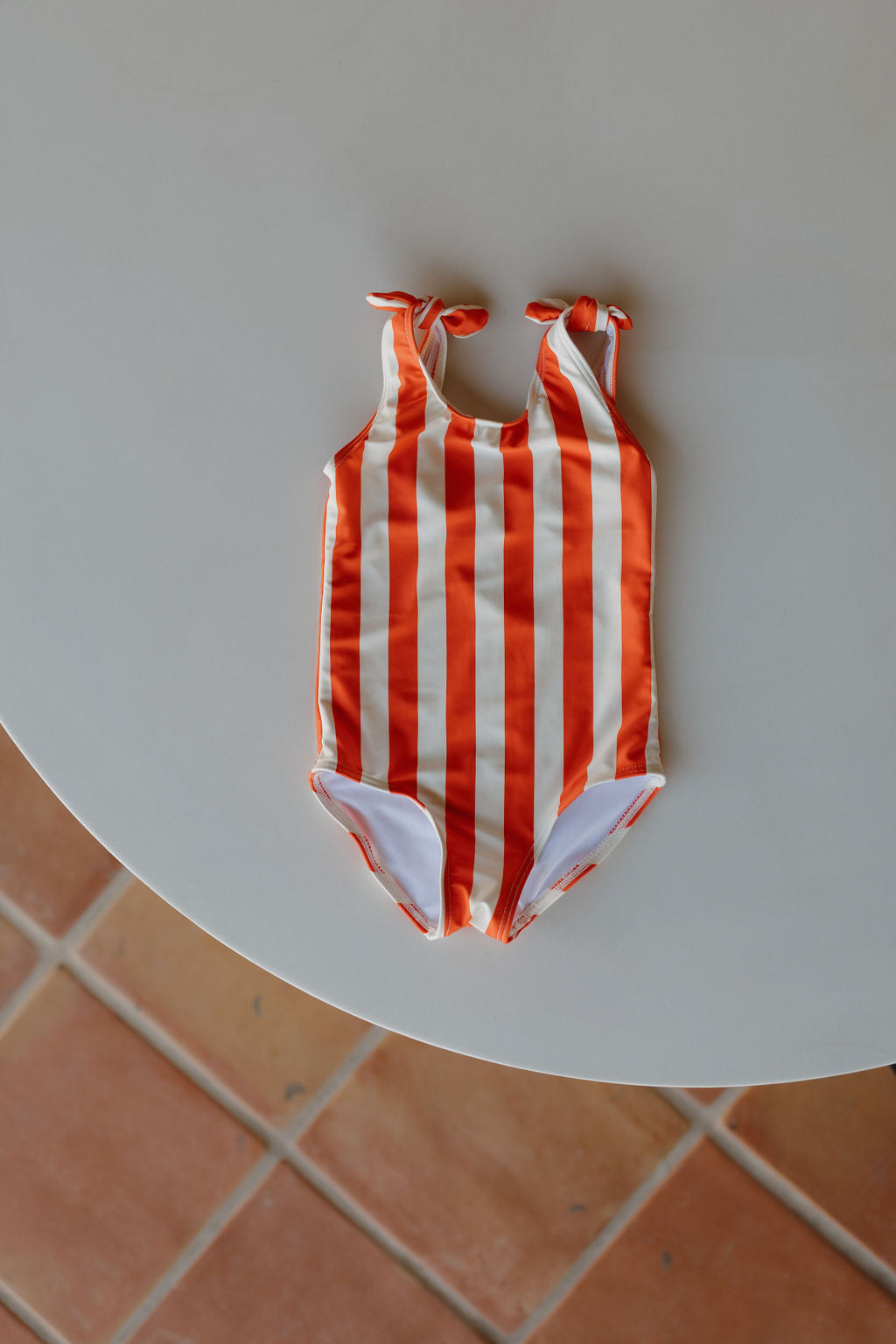 The Girls Sleeveless Swimsuit from the forever French baby Poolside collection, featuring red and white vertical stripes with knotted shoulder straps, is elegantly displayed on a round white table set on a tiled floor.