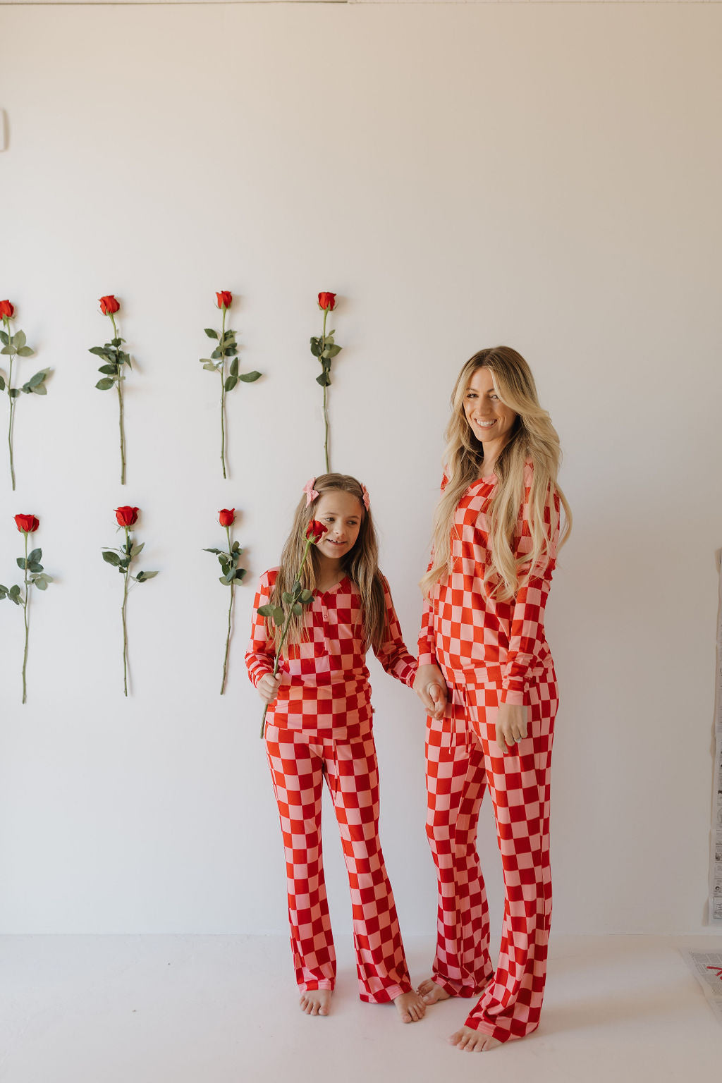 A woman and a young girl smile at the camera in matching Youth Flare Bamboo Pajamas by forever french baby, featuring a red and white checkered pattern. They are barefoot and holding hands, with roses arranged vertically on the white wall behind them.