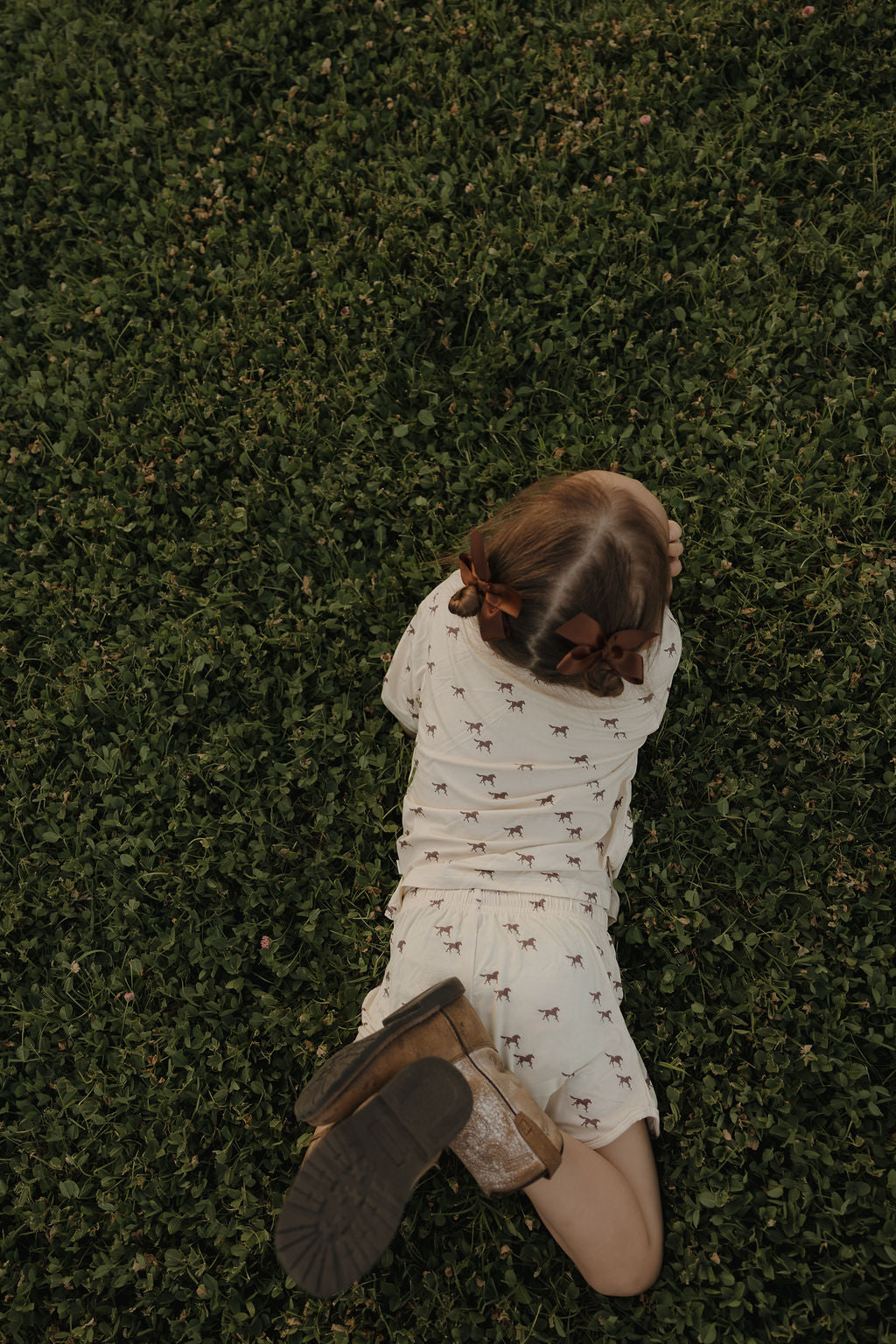 A young child with brown bows lies face down on green grass, wearing the Girl's Bamboo Short Set | Wild Horses by forever french baby and brown shoes.