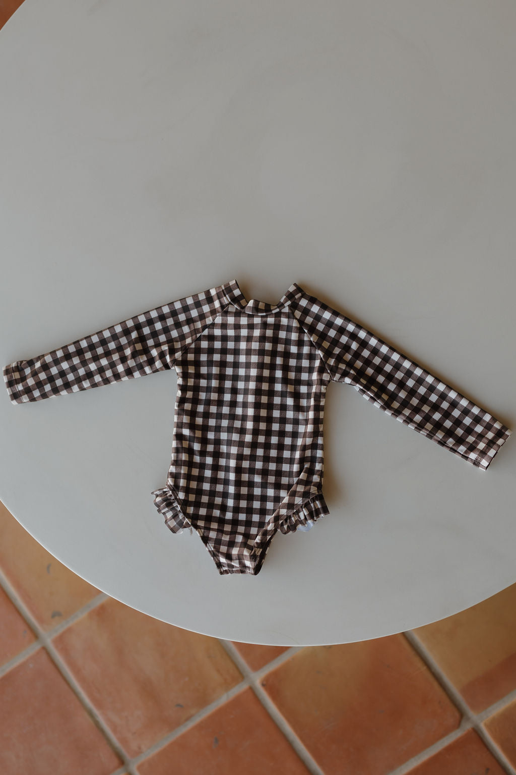 This delightful long-sleeve romper from forever French baby's Chocolate Gingham Collection features brown and white checks with ruffle details, elegantly displayed on a round white tabletop against a tiled floor.