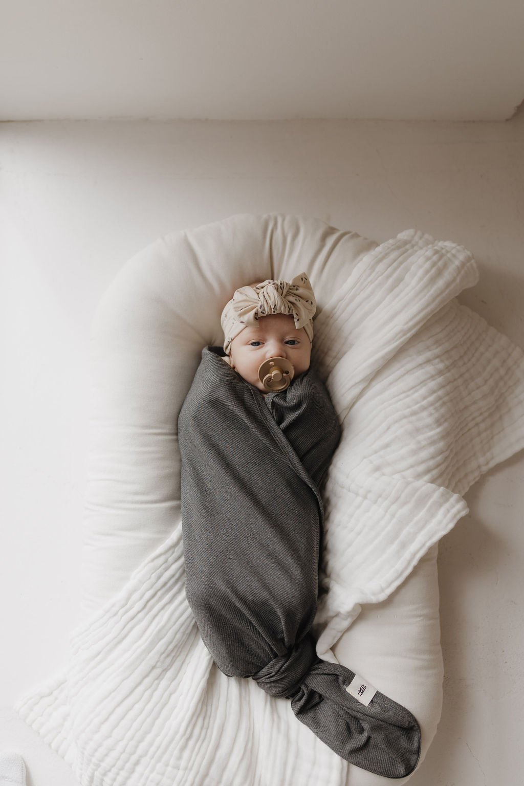 A baby wrapped in a forever french baby Bamboo Swaddle in Shadow lies on a white cushion, with a pacifier and wearing a light headband with a bow. Soft, natural light perfectly illuminates this hypoallergenic scene.