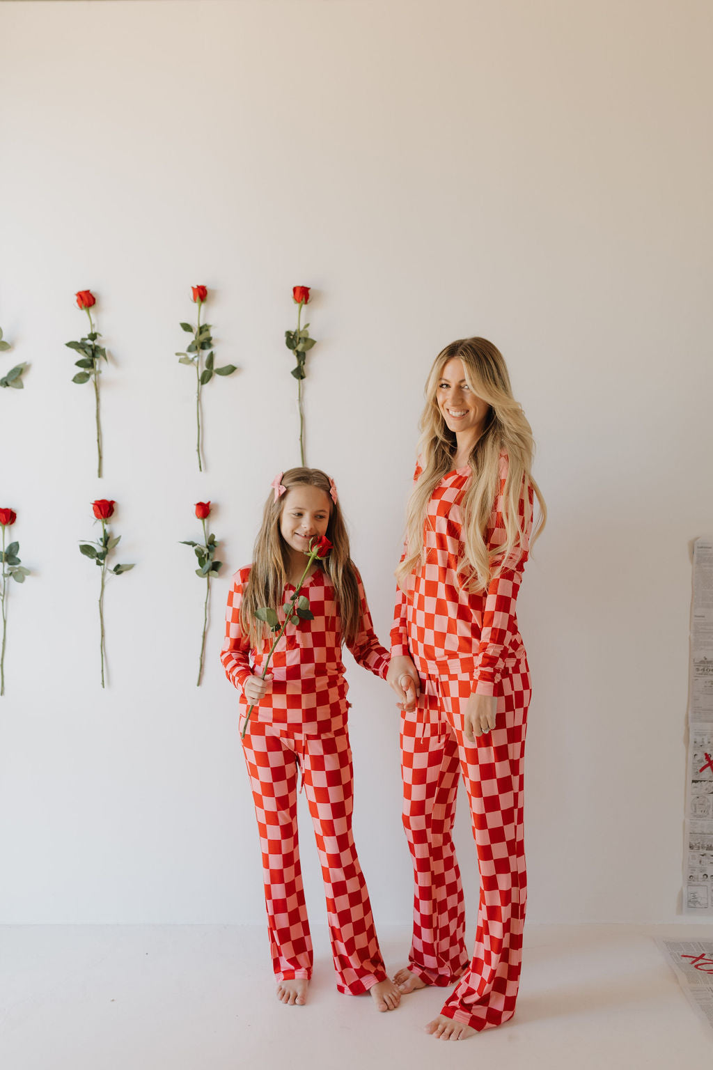 A woman and a young girl stand smiling, wearing matching "Youth Flare Bamboo Pajamas | XOXO" by forever french baby. They hold hands in front of a white wall adorned with red roses, the young girl holding a rose. Their cozy outfits exude comfort and style in red and white checkered fabric.