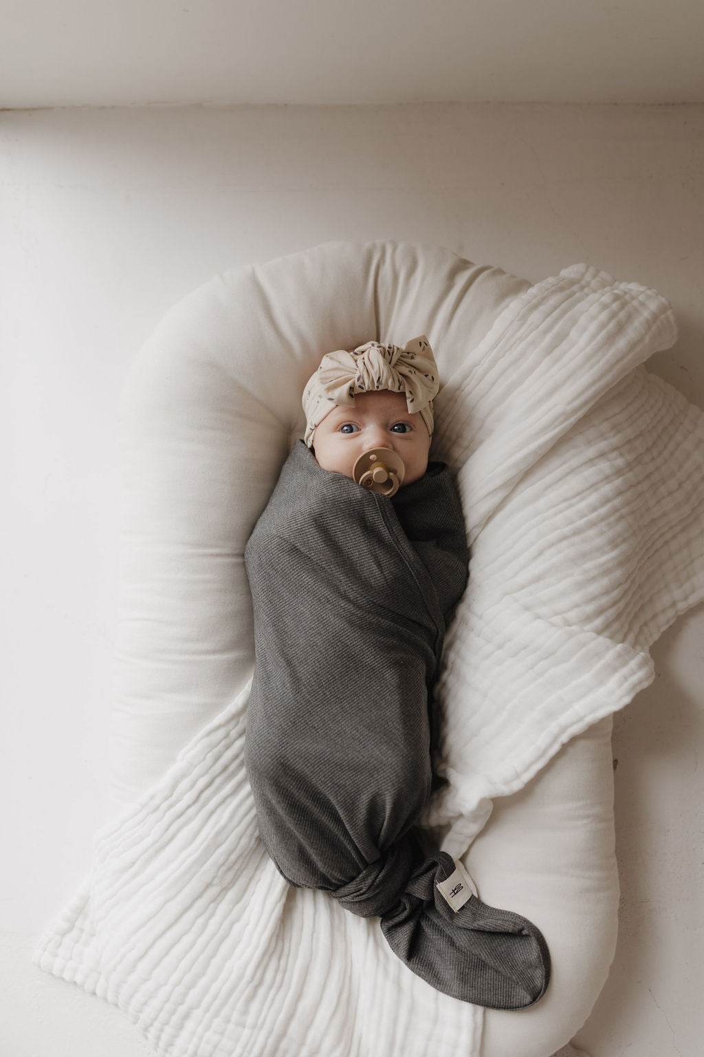 A baby peacefully rests on a white cushion, swaddled in the forever french baby's breathable hypo-allergenic Bamboo Swaddle | Shadow. The baby, with a patterned headband and pacifier, enjoys maximum comfort.