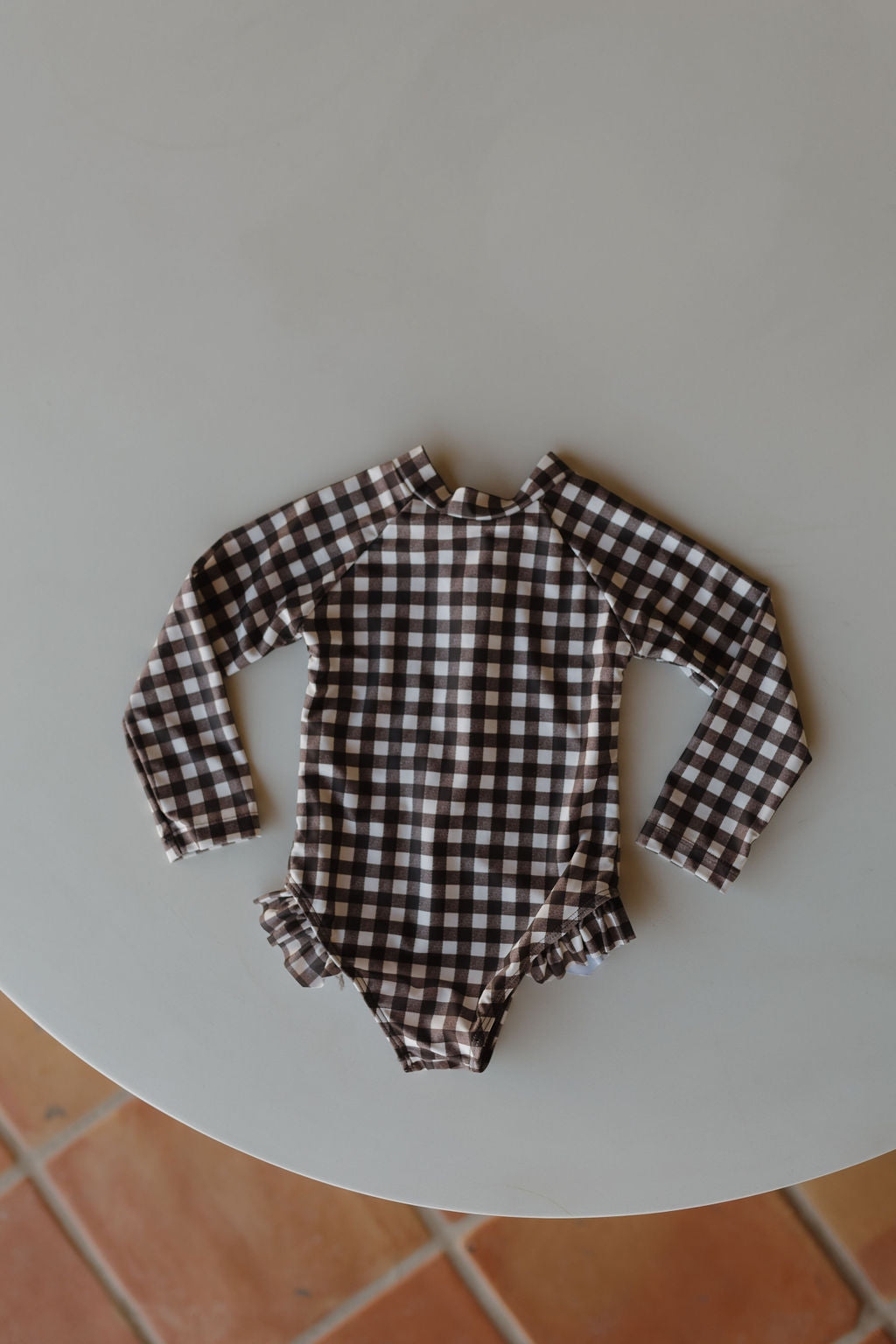 From the Forever French Baby collection, the Girls Long Sleeve Swimsuit in Chocolate Gingham is showcased on a light round table with terracotta tiles below, providing a warm contrast to this charming piece.