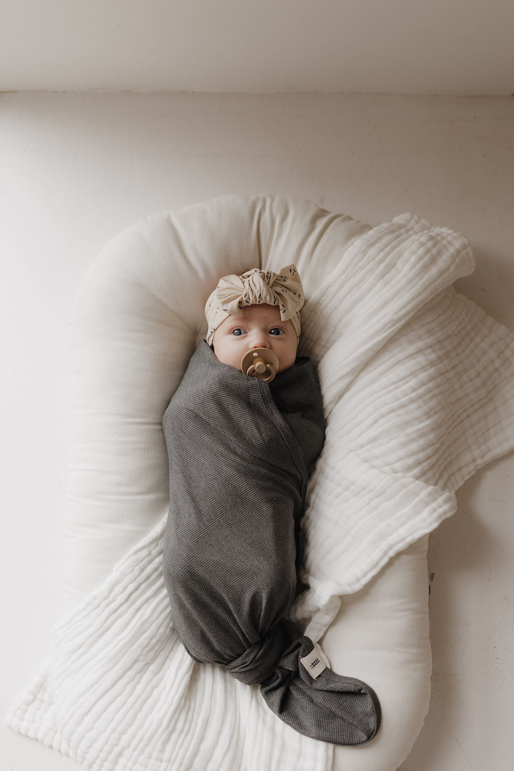 A baby is wrapped in a forever french baby Bamboo Swaddle in Shadow while lying on a white padded cushion. The hypoallergenic, breathable fabric ensures comfort. The baby has a pacifier and wears a patterned headband as soft, natural light illuminates the scene.