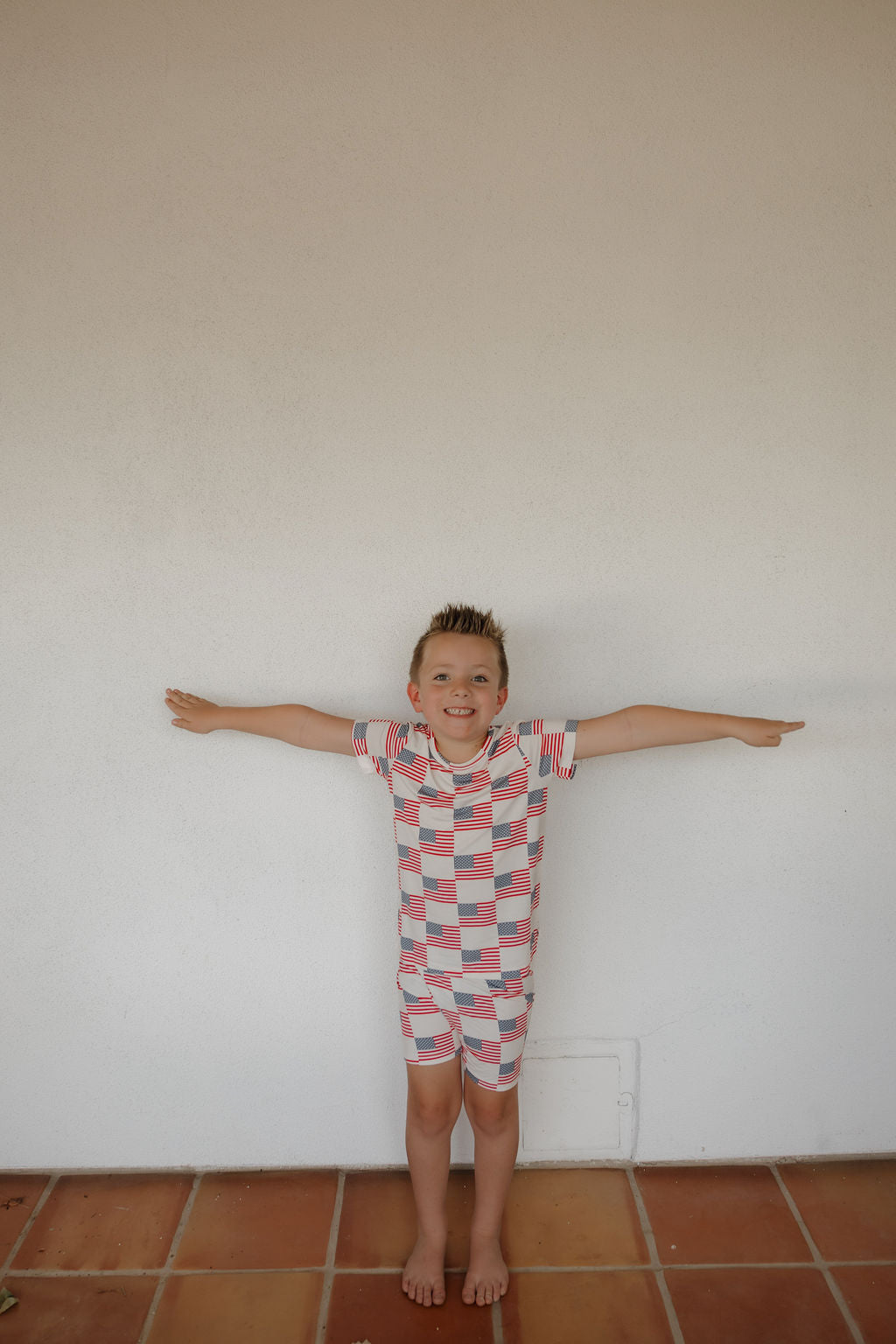 A young boy stands barefoot on a tiled floor against a plain white wall, smiling with his arms outstretched, wearing the forever french baby Bamboo Short Set | Americana made from breathable bamboo fabric.