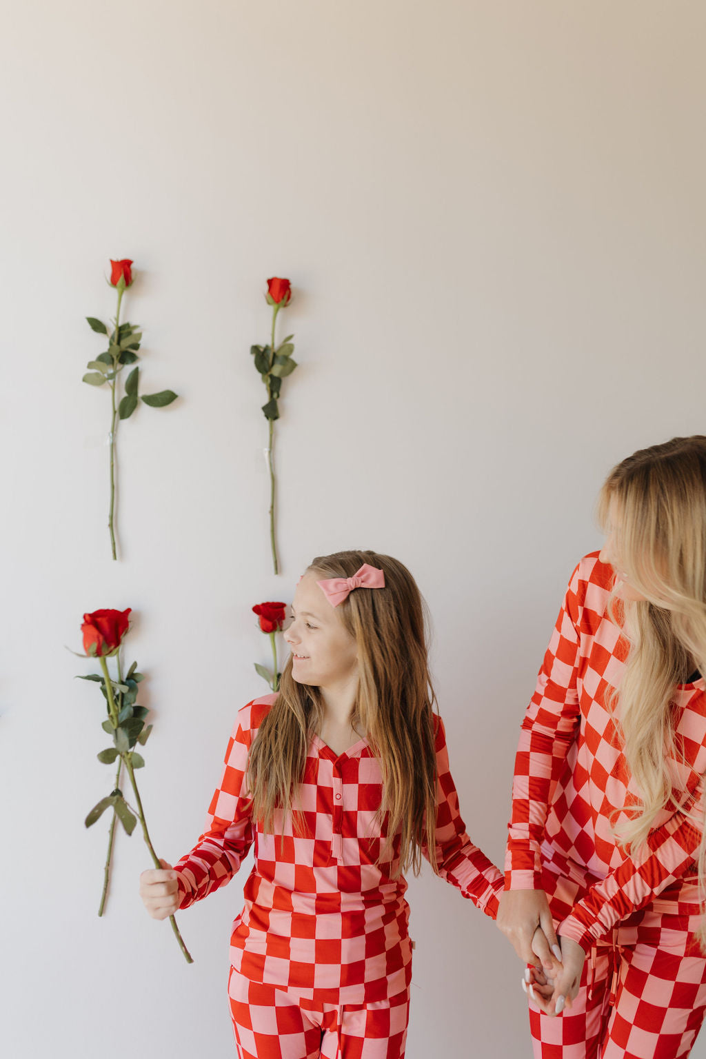 A child and an adult, both wearing matching Youth Flare Bamboo Pajamas by forever french baby in red and white checkered patterns, stand before a wall with red roses. The child holds a rose and smiles warmly at the adult.