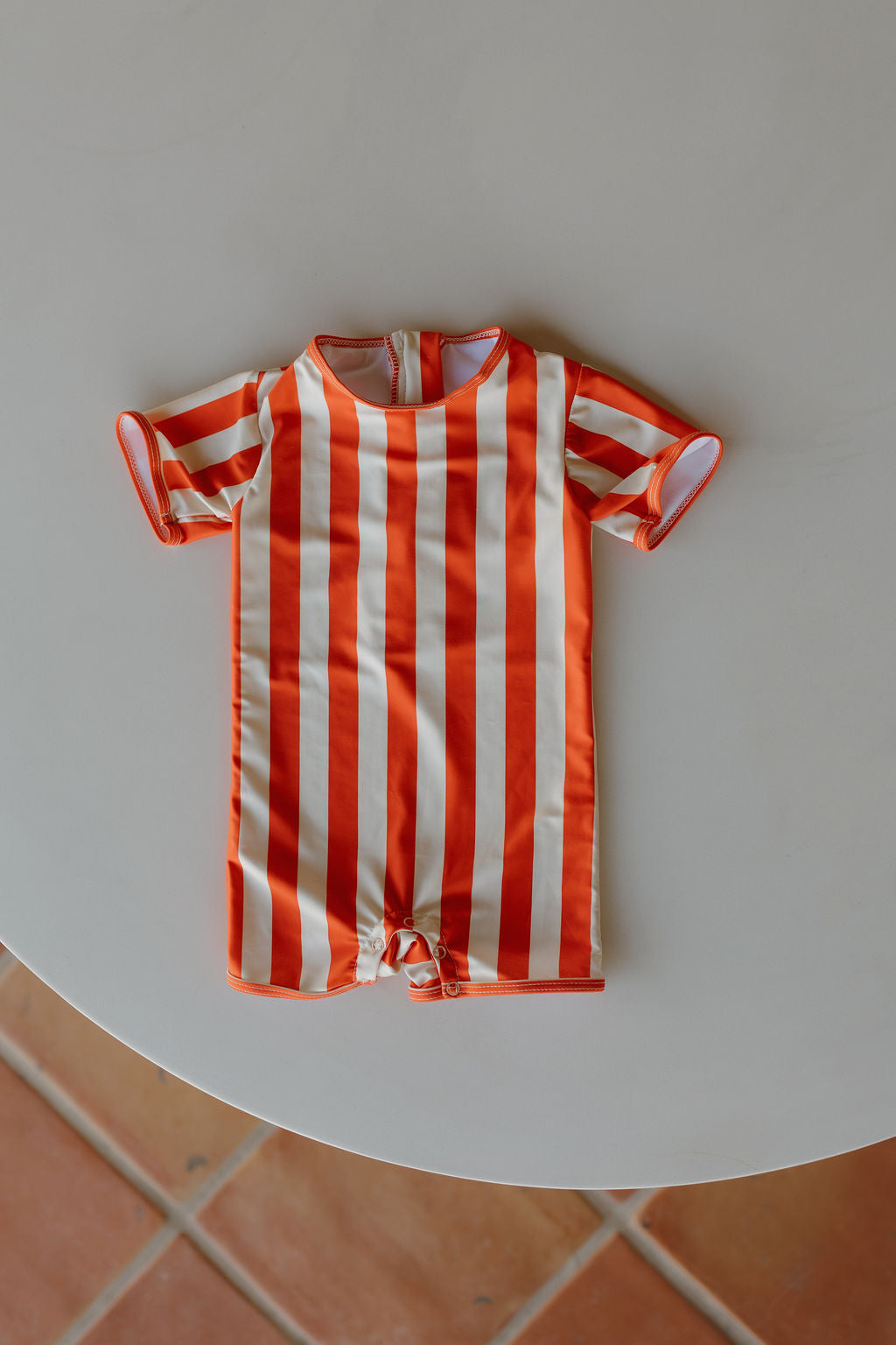 The Child Shortie Swimsuit from the Poolside Collection by forever french baby, adorned with vertical orange and white stripes, is elegantly presented on a round, white surface with a terracotta tile floor background.