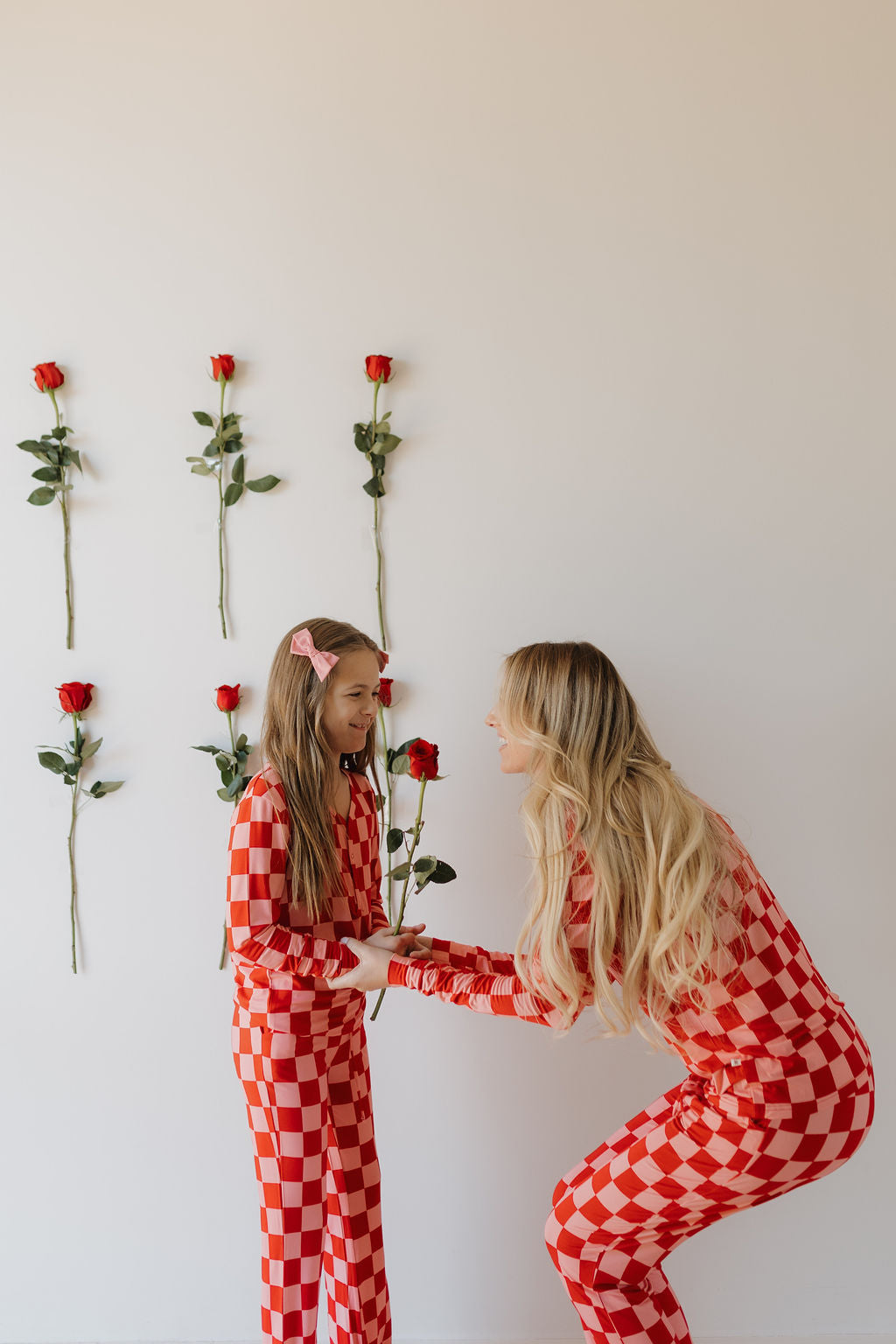 A woman and a young girl happily face each other in matching Youth Flare Bamboo Pajamas by forever french baby, showcasing red and white checkered designs. They stand in a white-walled room with vertical rows of red roses enhancing their joyful moment.