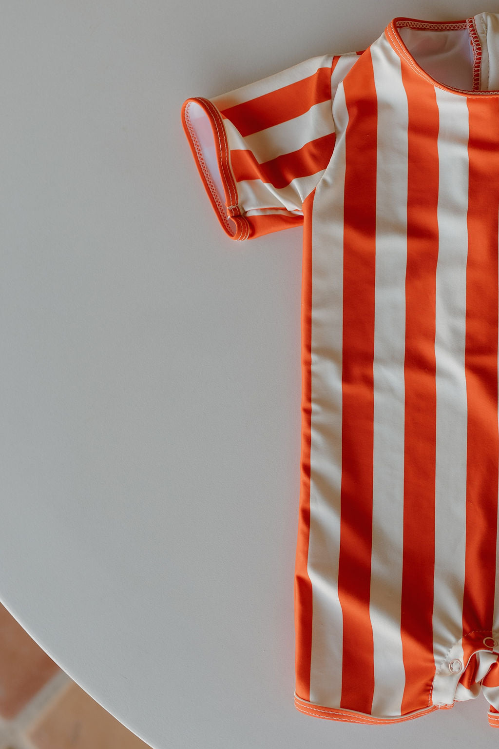 From the Poolside collection, the orange and white striped Child Shortie Swimsuit by forever french baby, featuring short sleeves, is displayed on a round white surface.