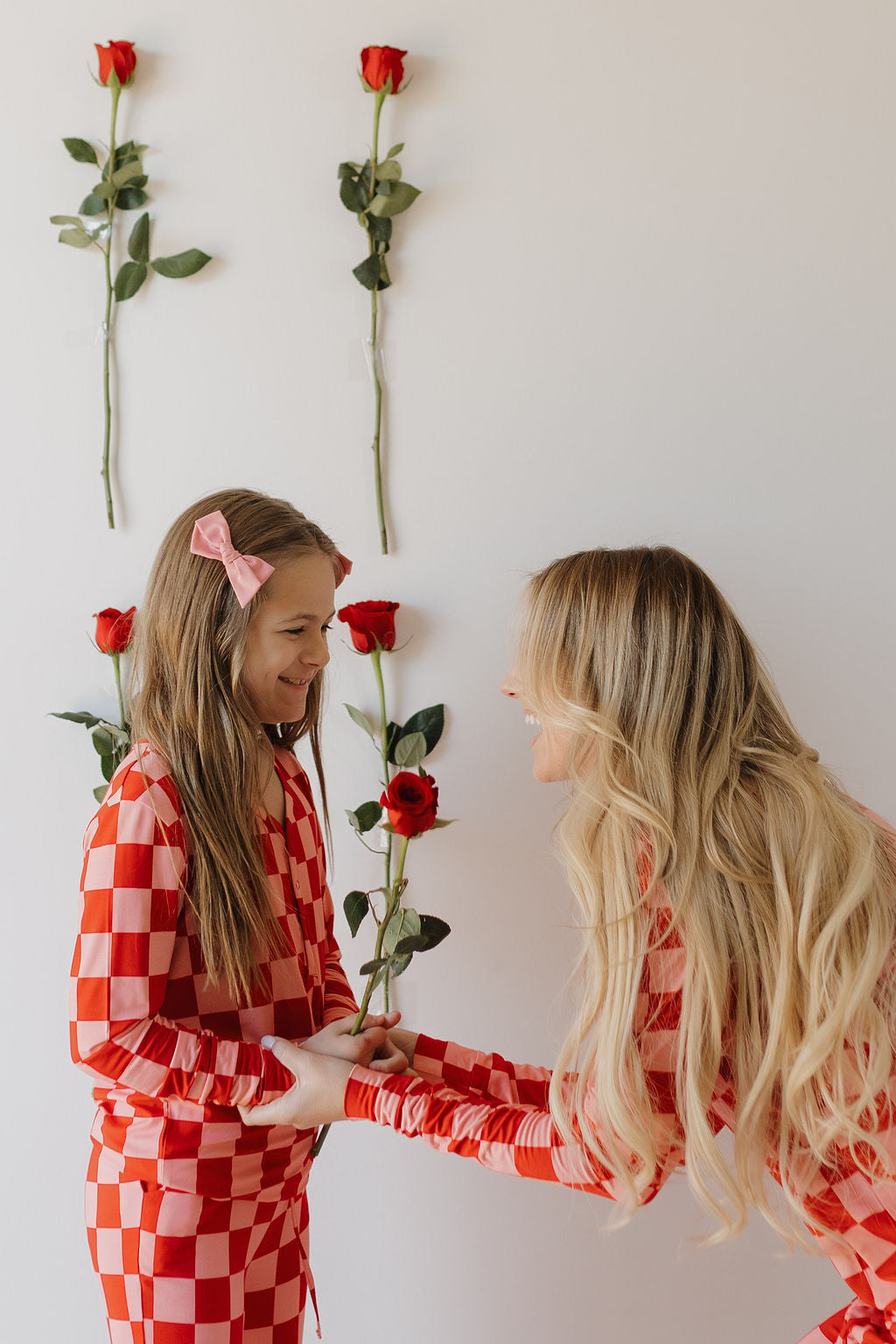 A woman and a young girl, both in matching Youth Flare Bamboo Pajamas by forever french baby, smile at each other. The woman leans toward the girl, holding her hands. Four red roses are vertically arranged on the white wall behind them.