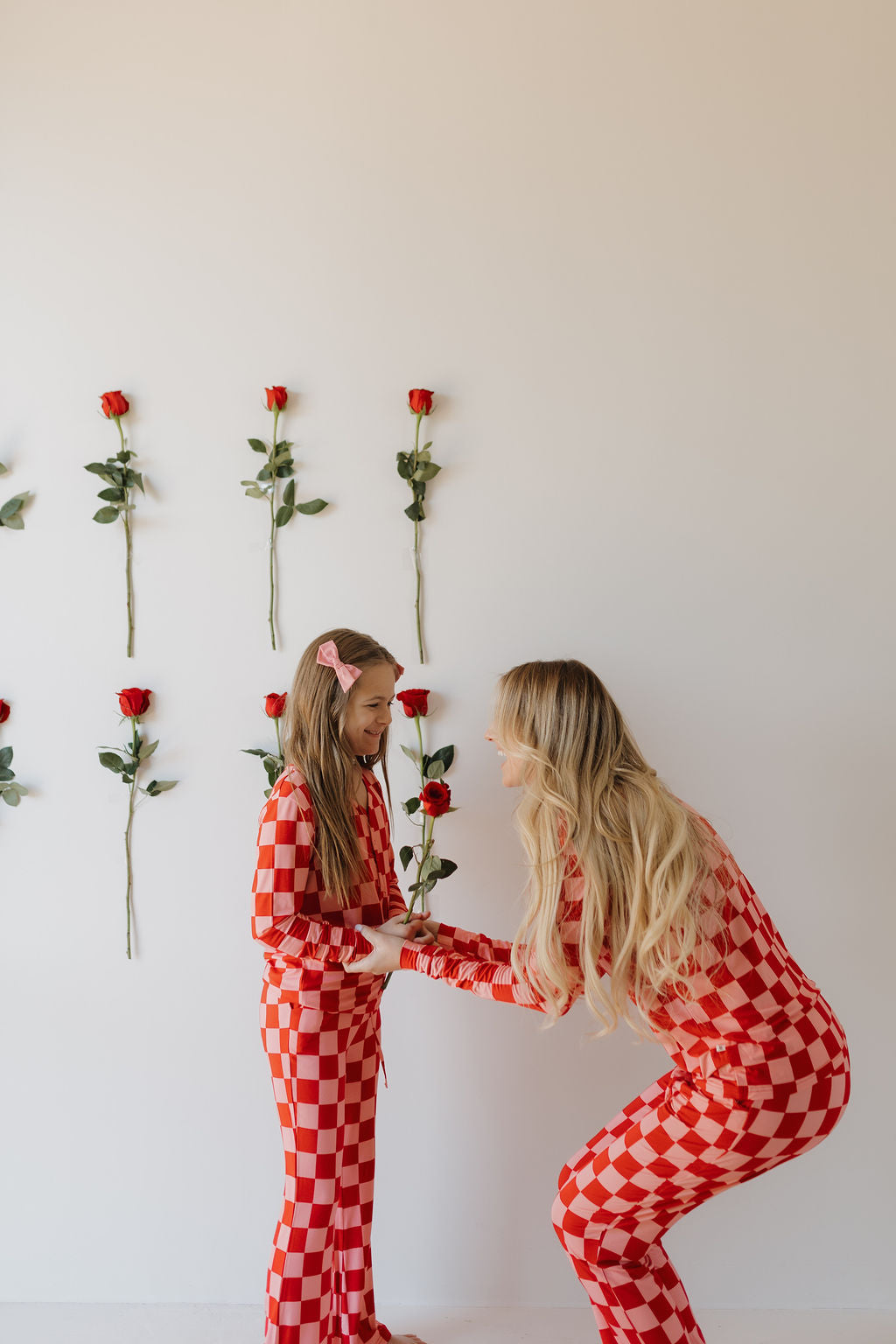 A woman and a girl in matching "Youth Flare Bamboo Pajamas | XOXO" by forever french baby stand before a white wall with vertical roses. The woman crouches, smiling at the girl, who wears a pink bow in her hair.