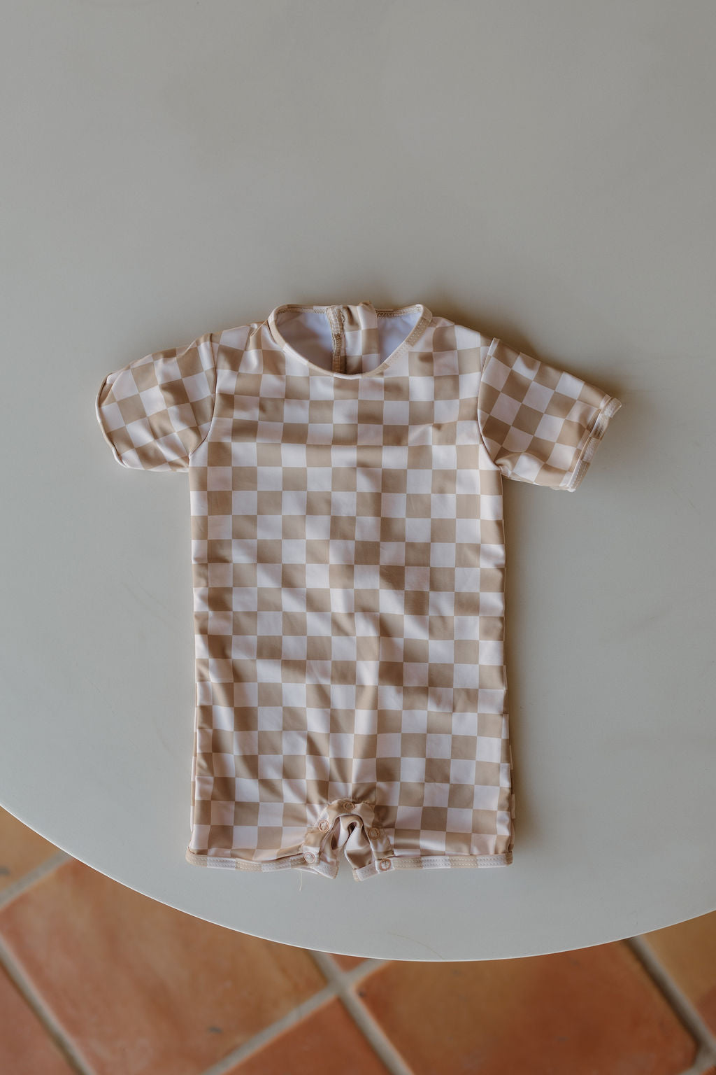 A Child Shortie Swimsuit from the Coastline Collection by forever french baby, showcasing a beige and white checkerboard design, is displayed flat on a white background from an overhead perspective.