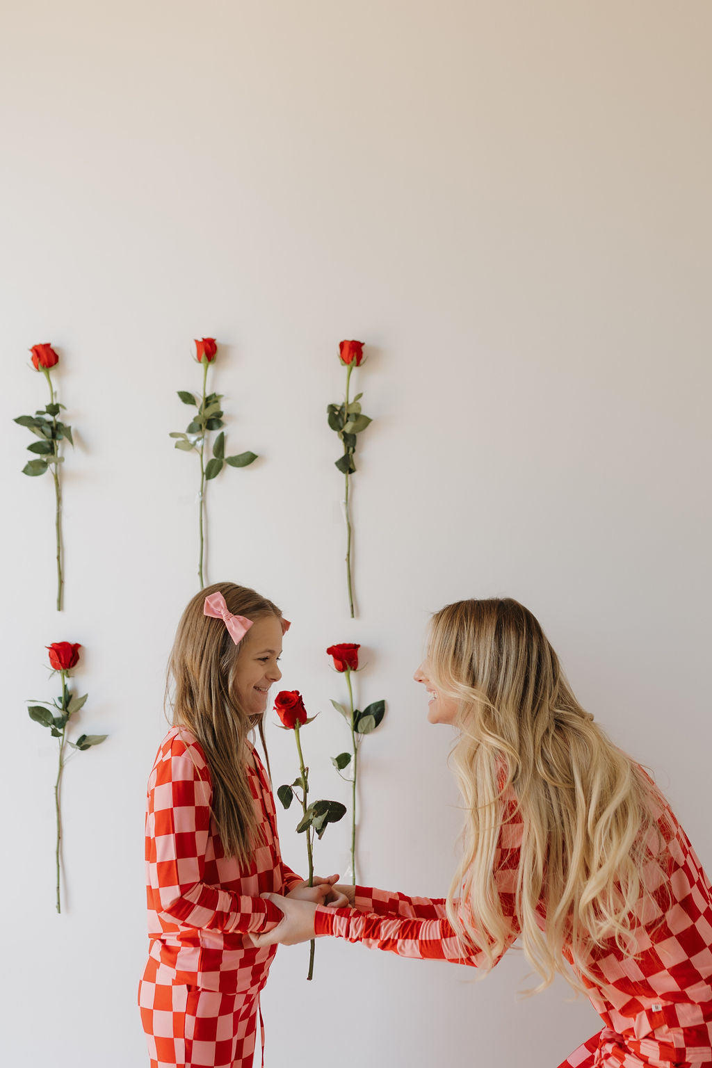 A woman and a girl are in red checkered outfits, with the girl in Youth Flare Bamboo Pajamas by forever french baby. They smile at each other while holding a rose, standing before a white wall decorated with six roses. The girl tops off her look with a pink hair bow.