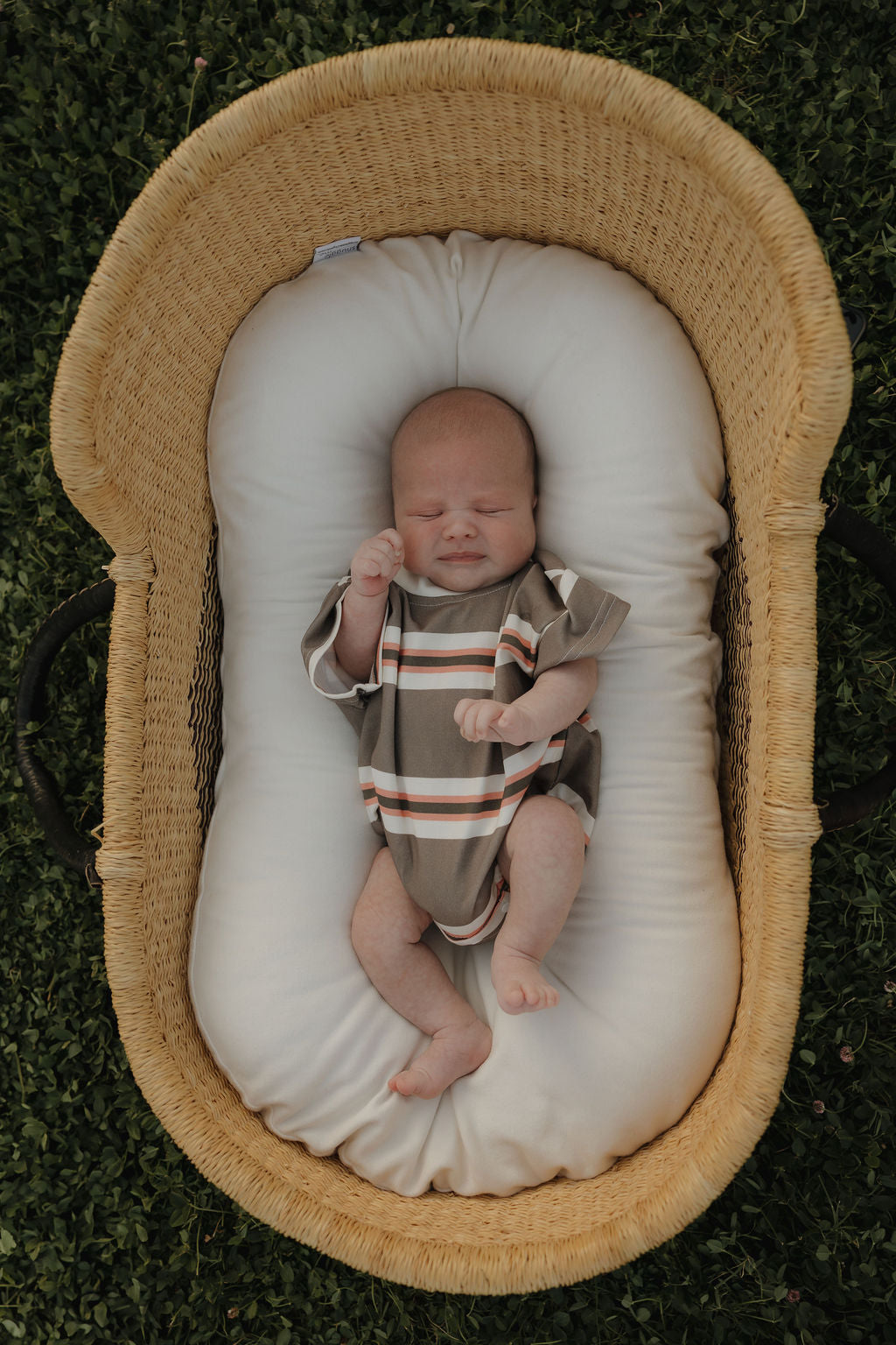 A newborn in the forever french baby Short Sleeve Romper | Sedona Stripe rests on a white cushion in a woven basket on green grass, eyes closed and hand near face—an adorable scene ideal for family matching outfits.