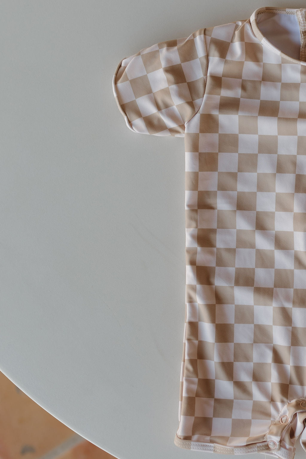 A Child Shortie Swimsuit | Coastline by forever french baby is laid out on a round white table, showing its checkered pattern with short sleeves and bottom snaps. The table edge and a glimpse of the floor are visible.