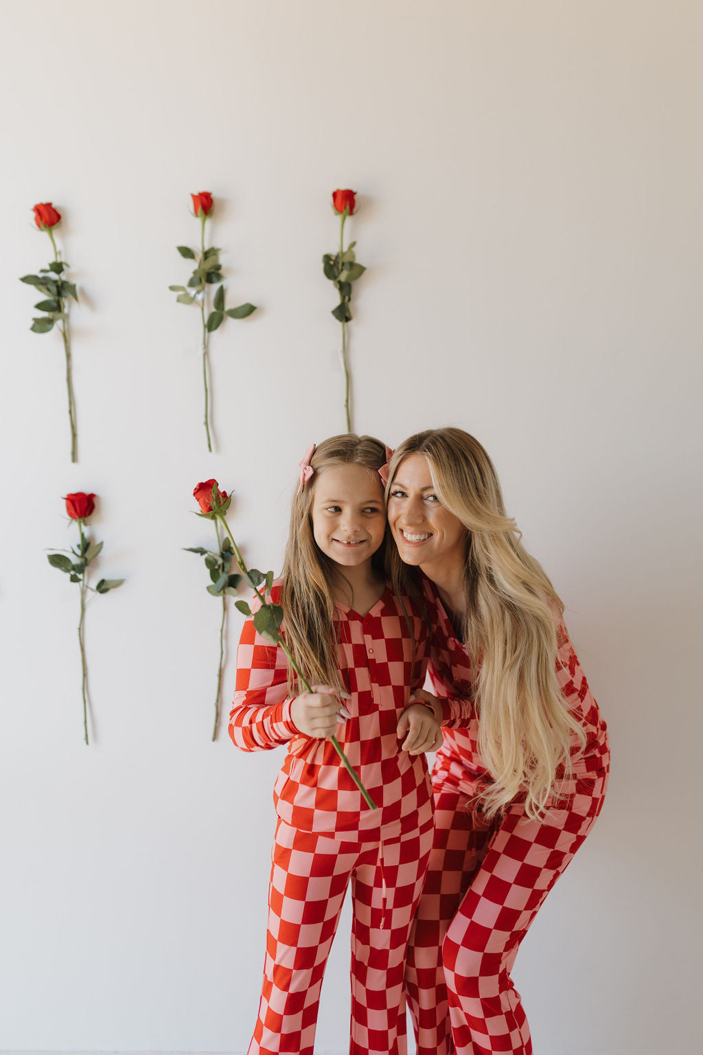 A woman and a girl with long hair wear Youth Flare Bamboo Pajamas from forever french baby, featuring red and white checkered patterns. They smile, each holding a rose, against a white wall decorated with six vertical roses.