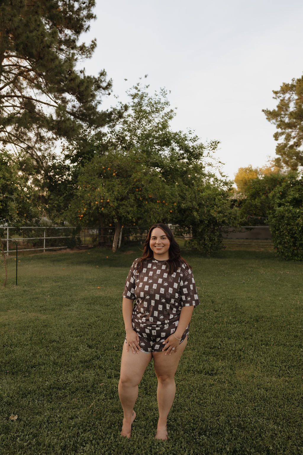 A woman stands barefoot on grass in a backyard, smiling and wearing the forever french baby Women’s Bamboo Short Set—hypo-allergenic and comfy. Trees and greenery fill the background under a partly cloudy sky.
