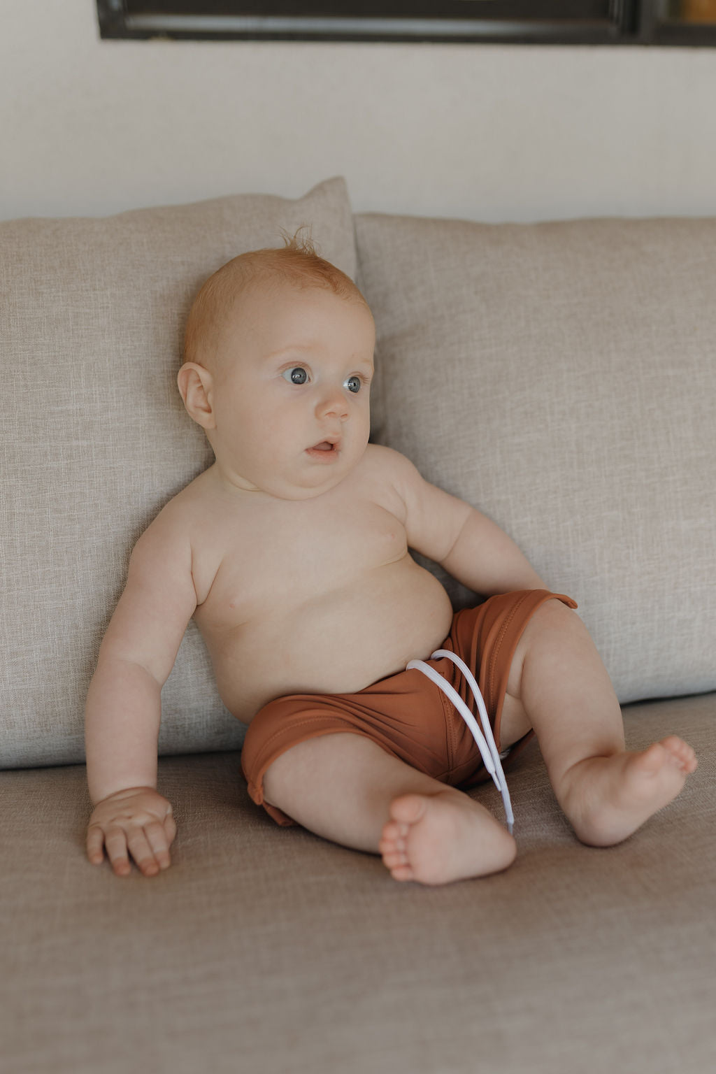 A light-skinned baby with short reddish hair sits on a beige couch, wearing the Terra collection's brown Child Boardshort by forever french baby. The shorts feature a white string for a customizable fit, and the baby gazes upward with a curious expression.