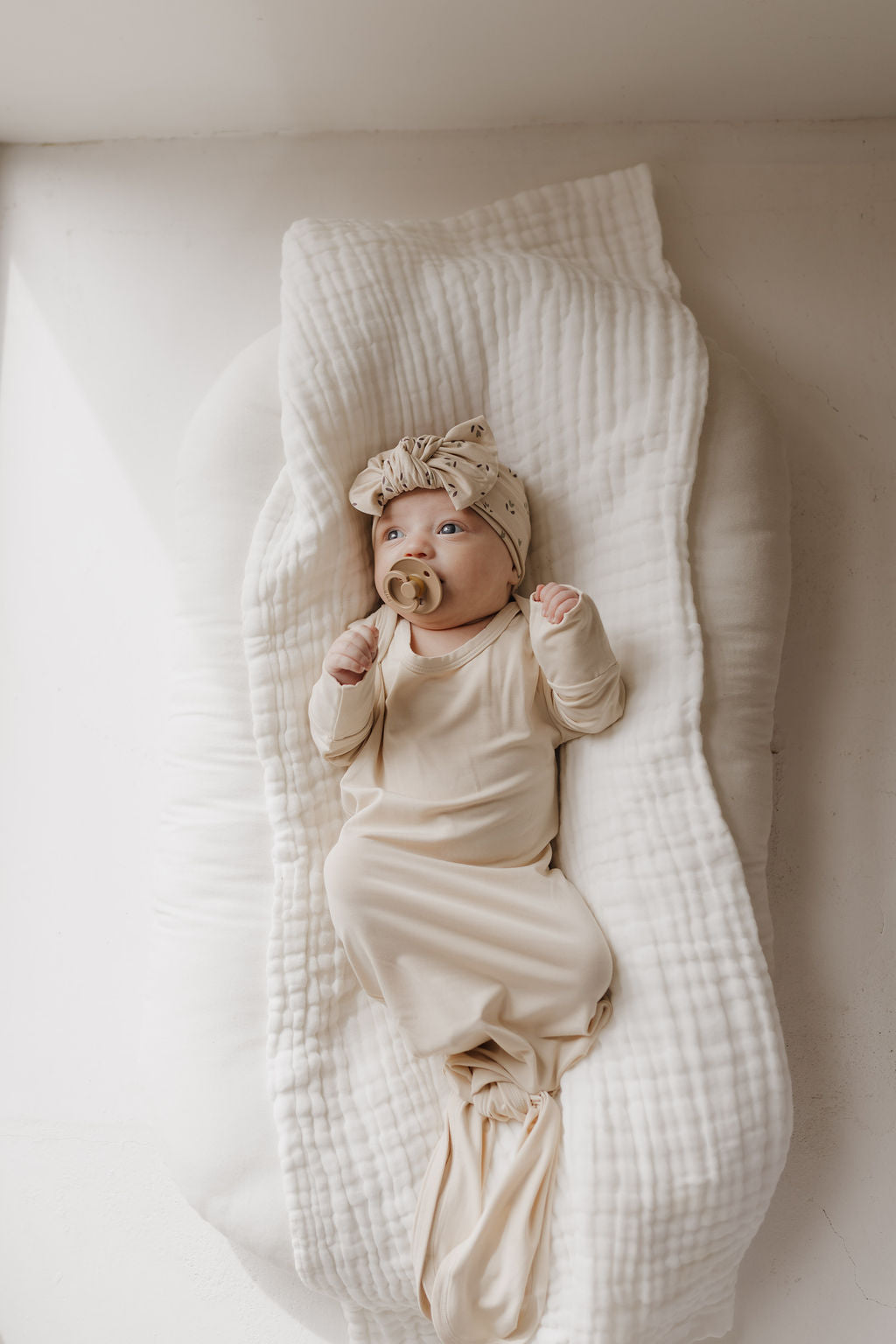 A newborn essential from forever french baby, the Bamboo Knotted Gown in Bone and matching headband are crafted from hypo-allergenic bamboo. The infant lays on a soft, white cushion with a pacifier, gazing upward in gentle natural light.